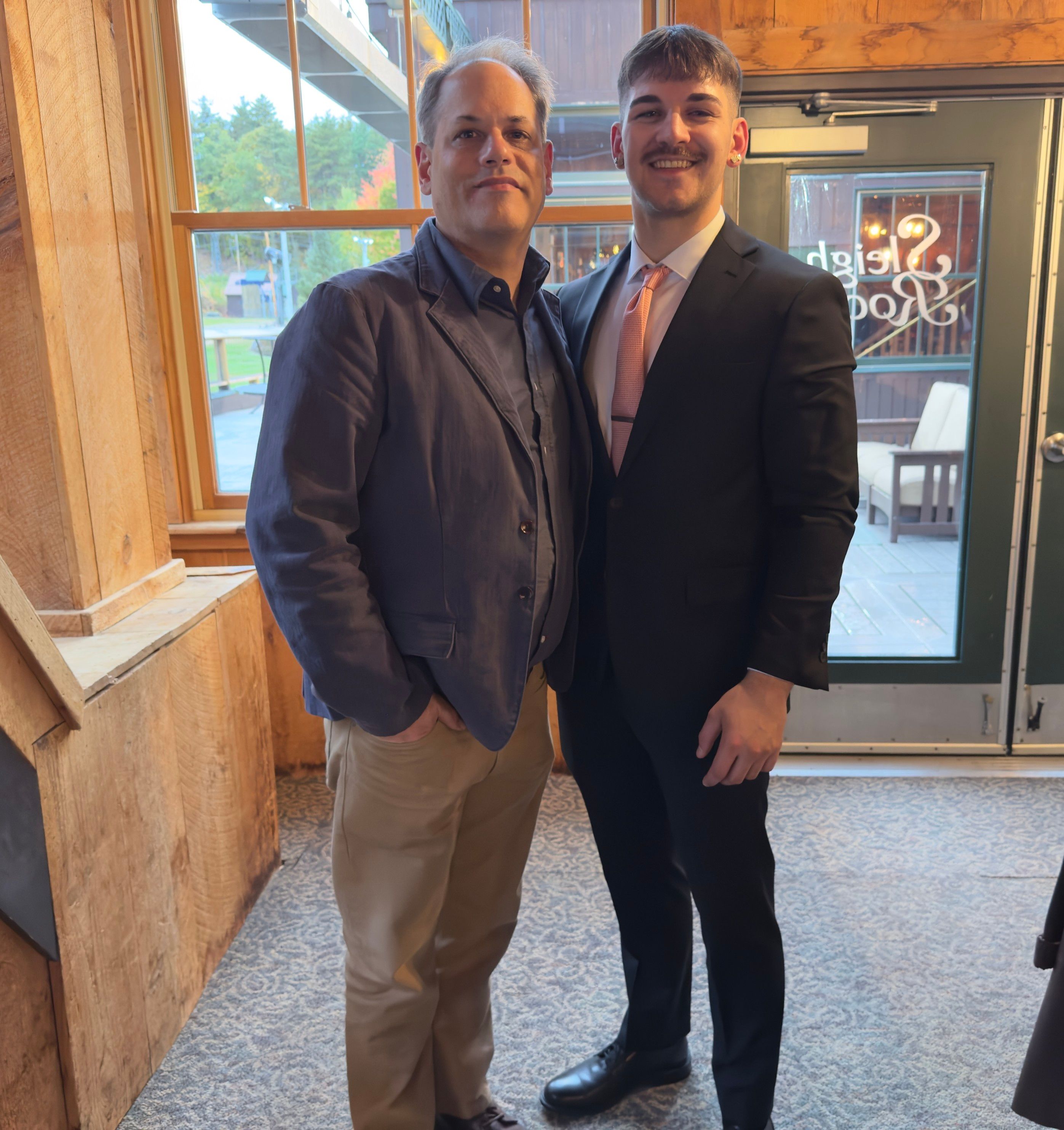 Two men are posing for a photo indoors. One man, wearing a blue jacket and beige pants, stands casually with his hands in his pockets. The other man is dressed in a black suit with a light pink tie. They are father and son. 