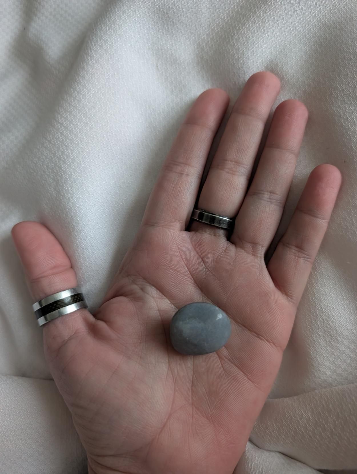 A photo of my left hand, laying open on a white bedspread, palm up. I have silver rings on my thumb and middle finger. A grey polished rock sits in the center of my open palm.