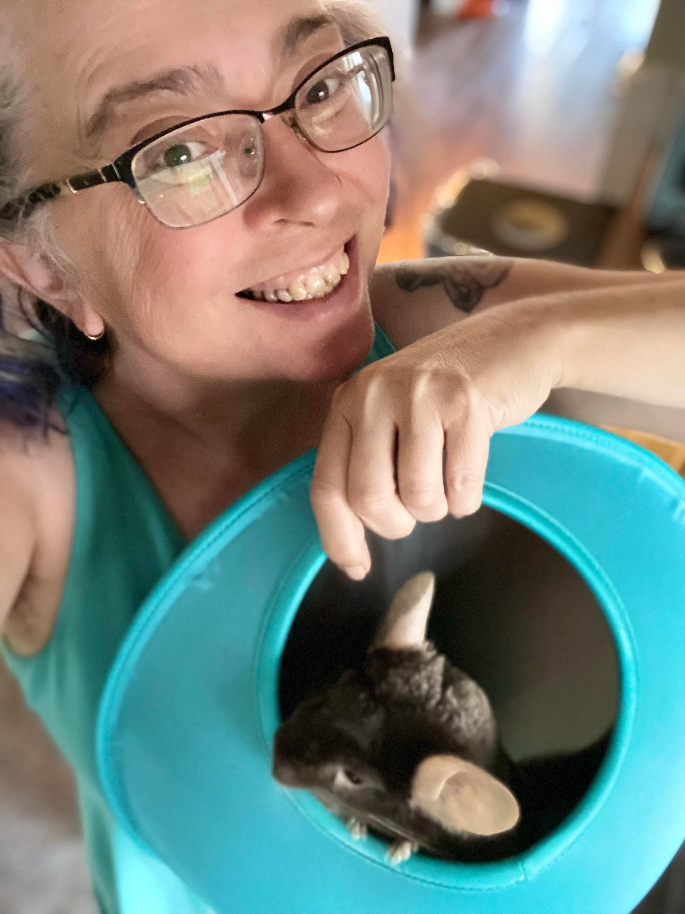 A smiling person with glasses holds a soft turquoise container, peering into it. Inside the container, a chinchilla can be seen, partially emerging.
