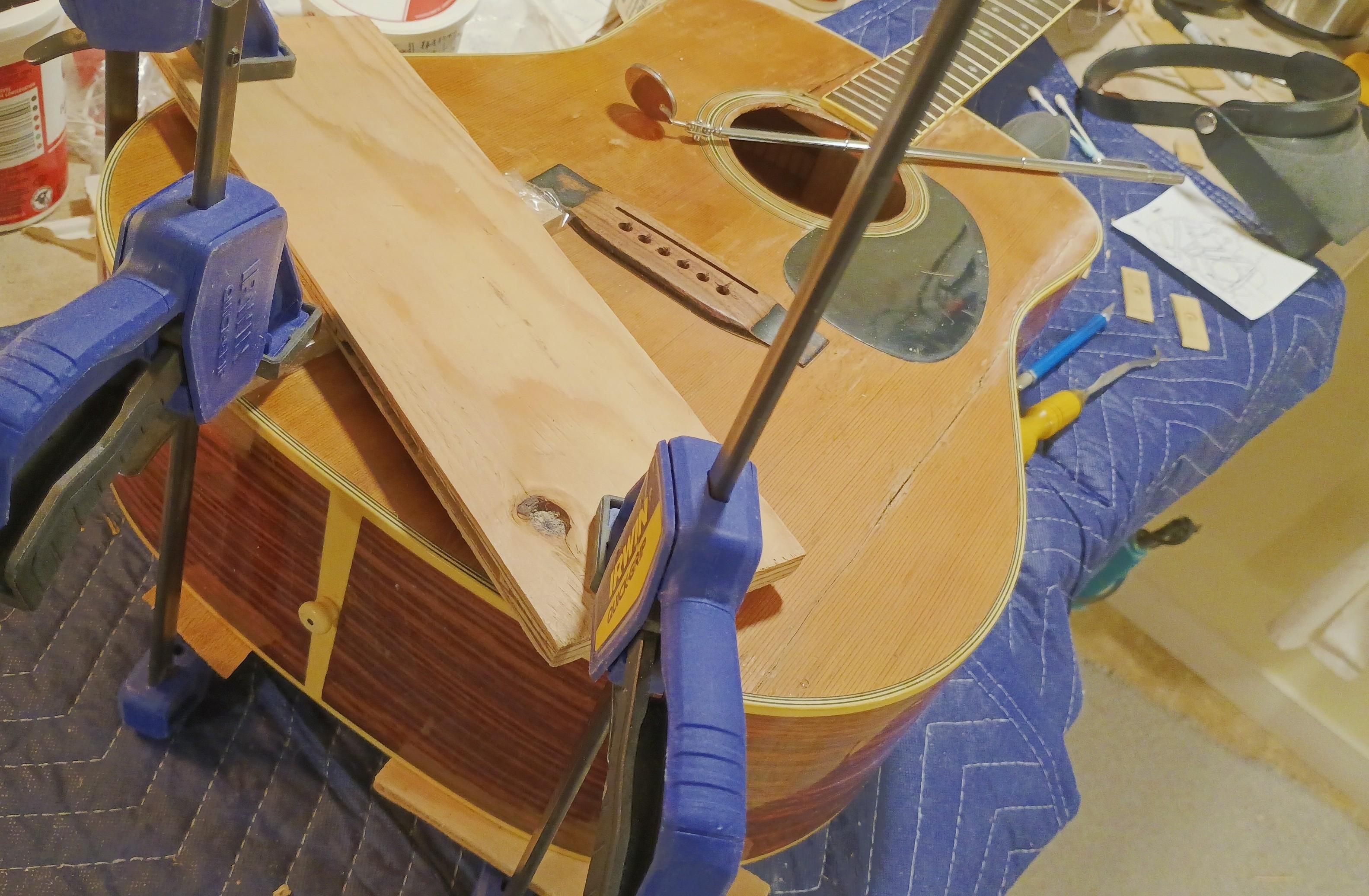 A guitar in a workbench sits on a padded moving blanket and three clamps good scrap wood in place while glue sets.