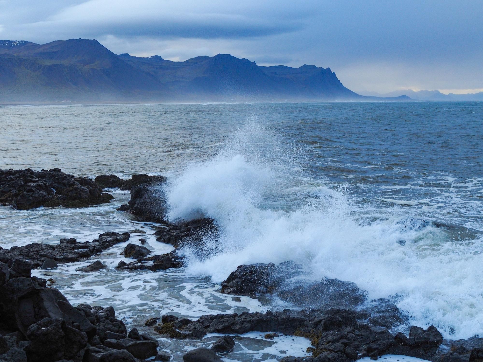 Waves crashing on black, rocky outcrop on a dark, moody, stormy day. A mountain range can be seen in the background. 