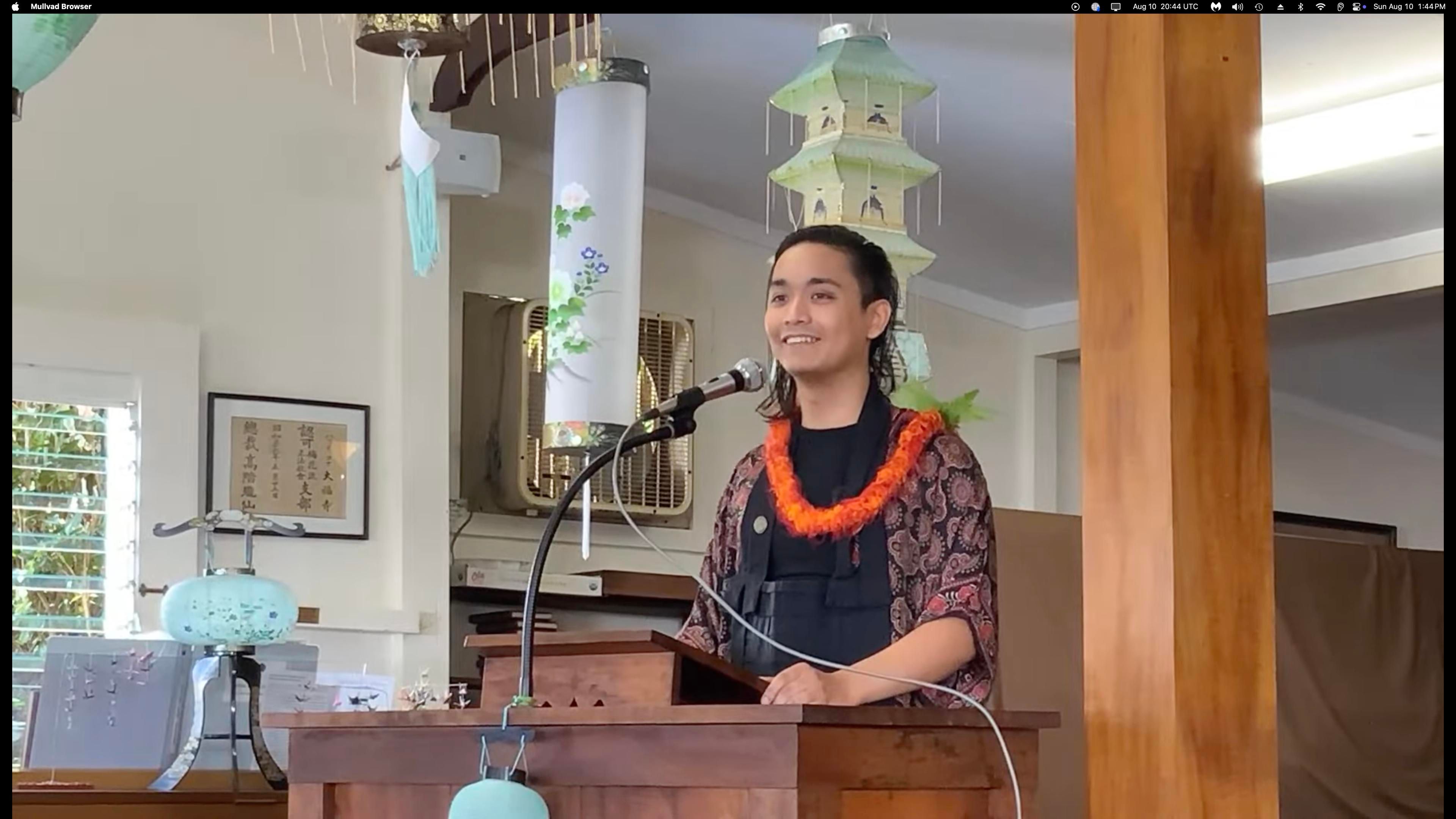 The talk “Waking Up in Challenging Times” was given by Daifukuji member Jordon “Soujin” Phillips. Soujin is a trans writer, editor, activist, and Zen student who is presently pursuing chaplaincy training within the Buddhist tradition and while working at Hospice Of Kona. The picture shows a woman wearing a bright lei speaking from a podium. 