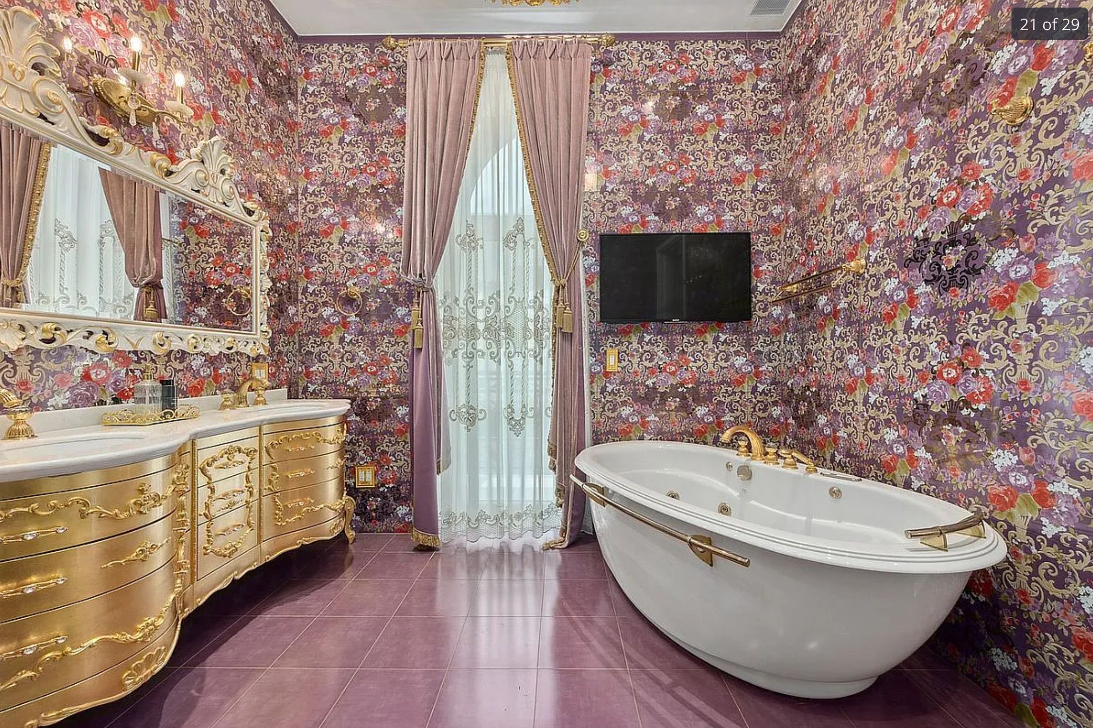 An utterly garish bathroom with very busy purple floral wallpaper, a purple tiled floor, purple curtains, a gold vanity with ornate mirror, a free standing bath and a wall mounted TV