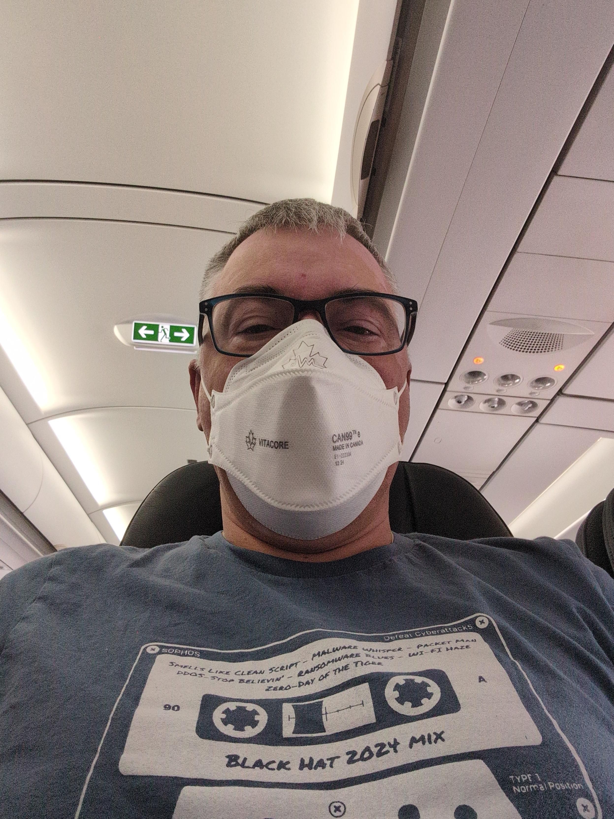 A picture of the author, sitting in a coach seat on Air Canada wearing a face mask and looking unimpressed. 