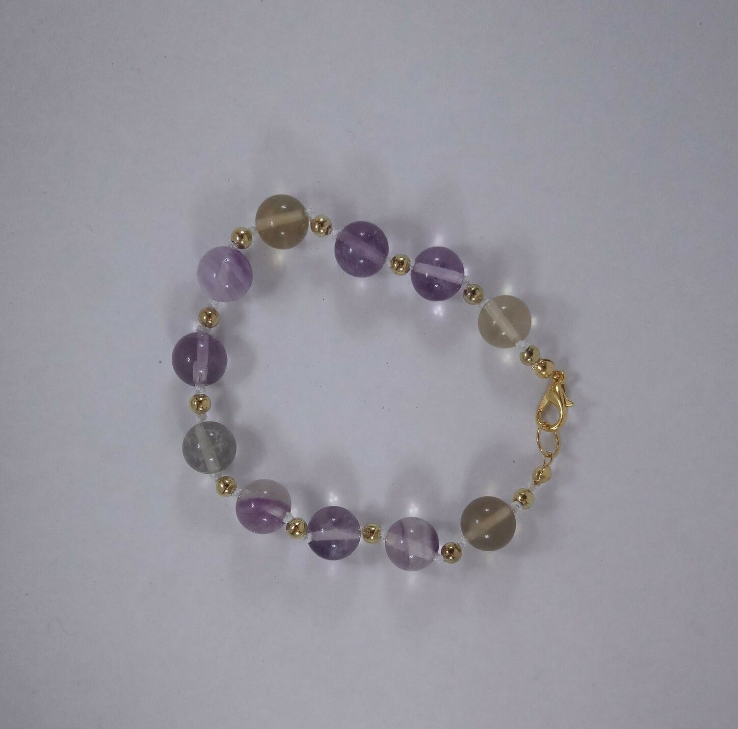 A fluorite bead bracelet, strung on white nylon cord with gold spacer beads. The fluorite beads range from pale purple to pale yellow, and they're transparent as well.