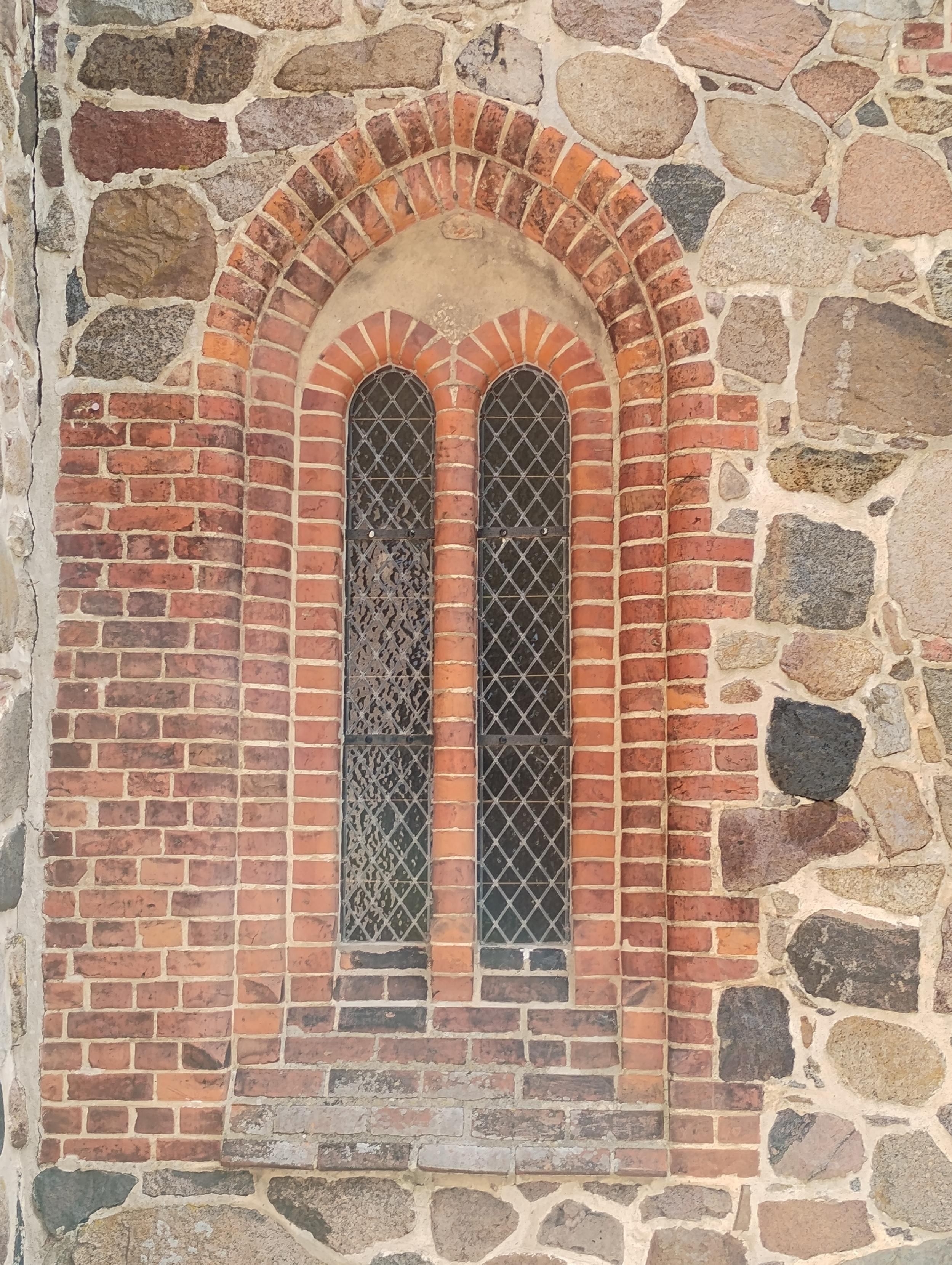 Ein gotisches Kirchenfenster, Mauerwerk aus rotbraunen Ziegeln, die Wand daneben aus bunten Findlingen gemauert.