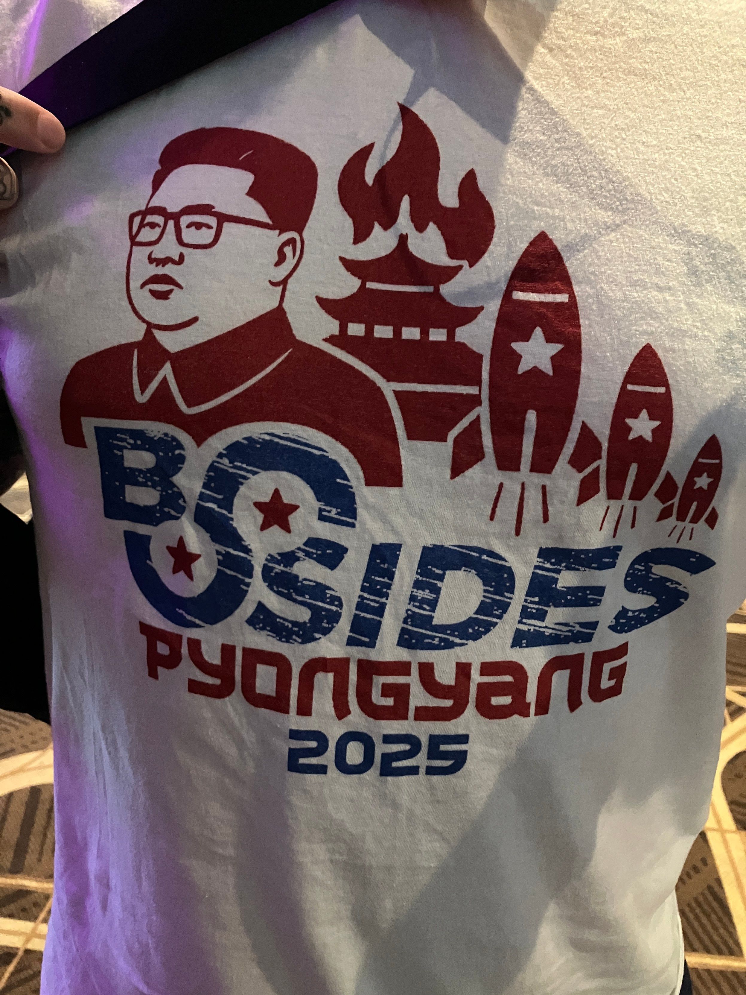Photo of a white T-shirt that has a picture of Kim Jong-Il and says BSides Pyongyang 2025