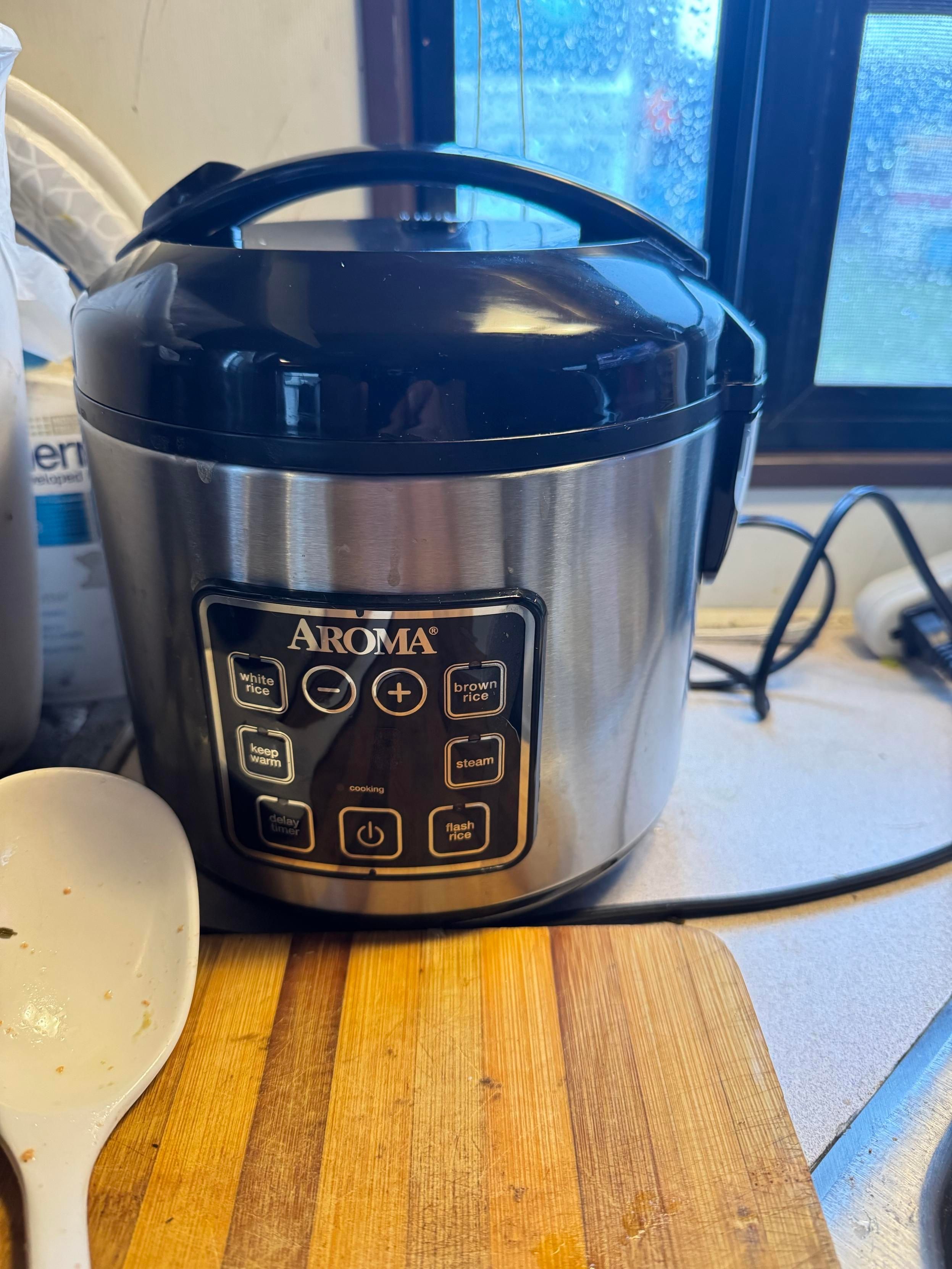An Aroma rice cooker is positioned on a countertop, featuring a black lid and a stainless steel body. The control panel has buttons for cooking white and brown rice, keeping warm, steaming, and a delay timer. A wooden cutting board 