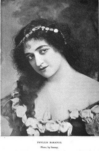 Phyllis Barasco, from an 1897 publication; a white woman with long dark curly hair, wearing a low-cut dress with flowers along the neckline, and a jeweled hair ornament