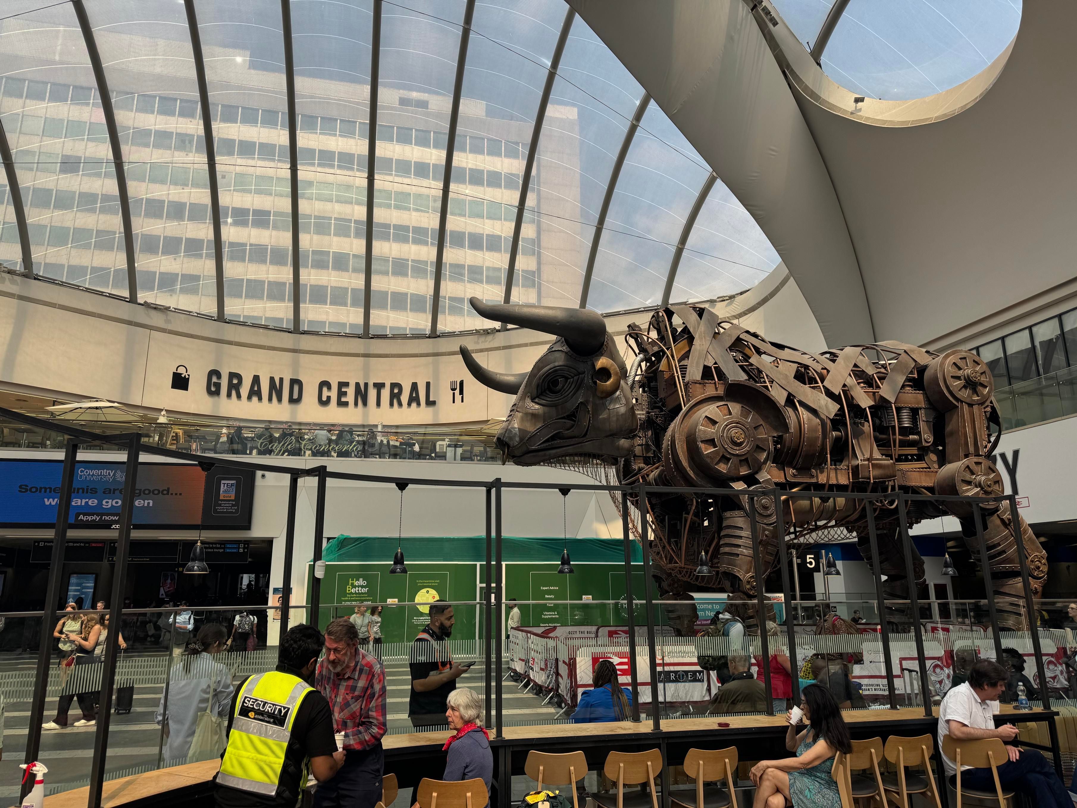View of Ozzy the Bull, a large bull made out of iron, in a large shopping centre that says “Grand Central” on the wall on the floor above. A large glass ceiling lets natural light in.