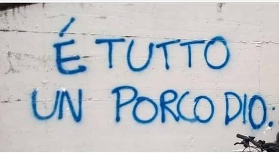 Una scritta a spray blu su un muro bianco: "E' tutto un porco dio".