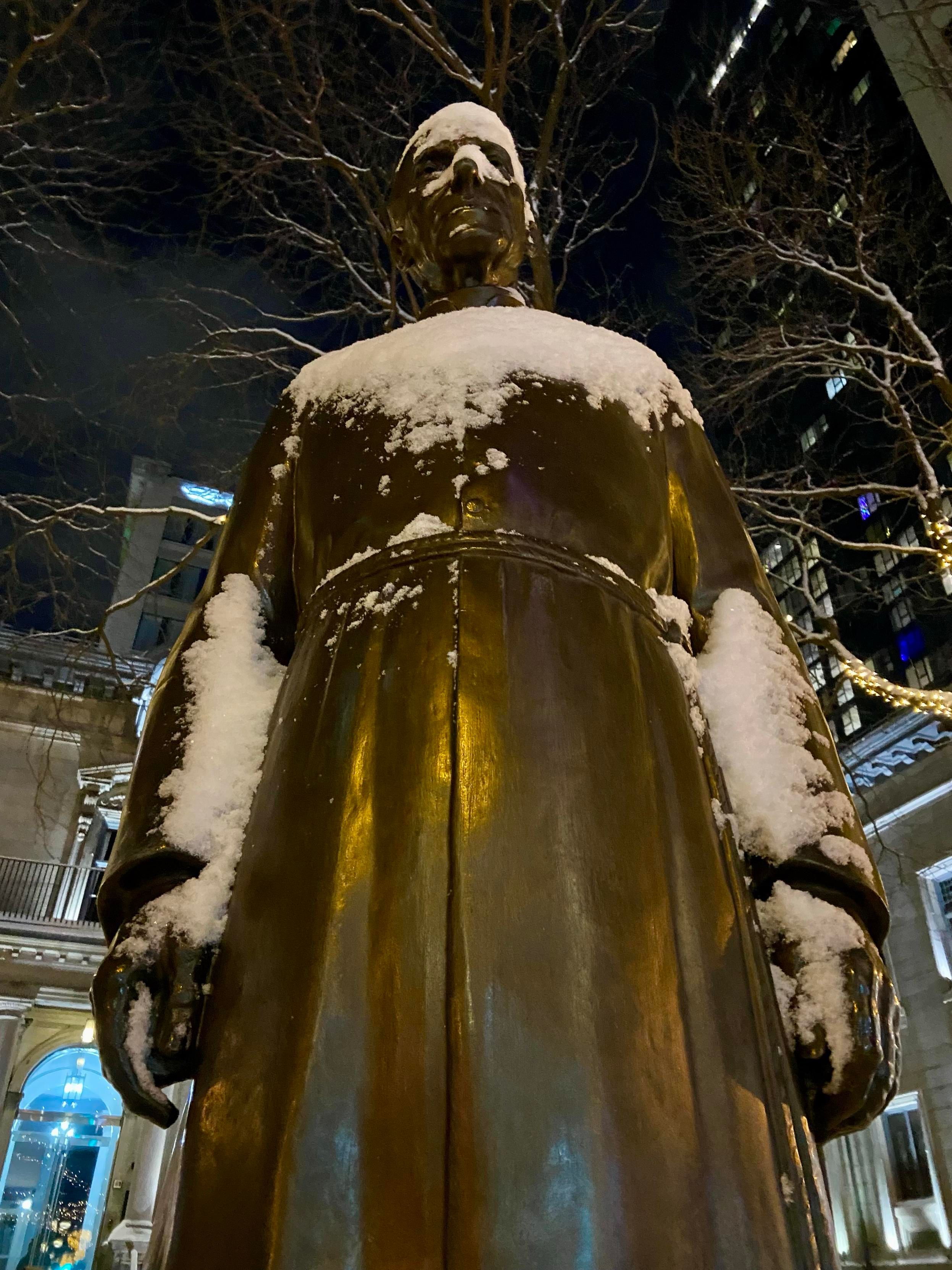 Looking up from the base of a bronze statue of frère André, on a snowy night. There's a light covering of snow on his head and shoulders and forearms. And just enough snow accumulated between his lips that it looks he's baring his teeth at us. 