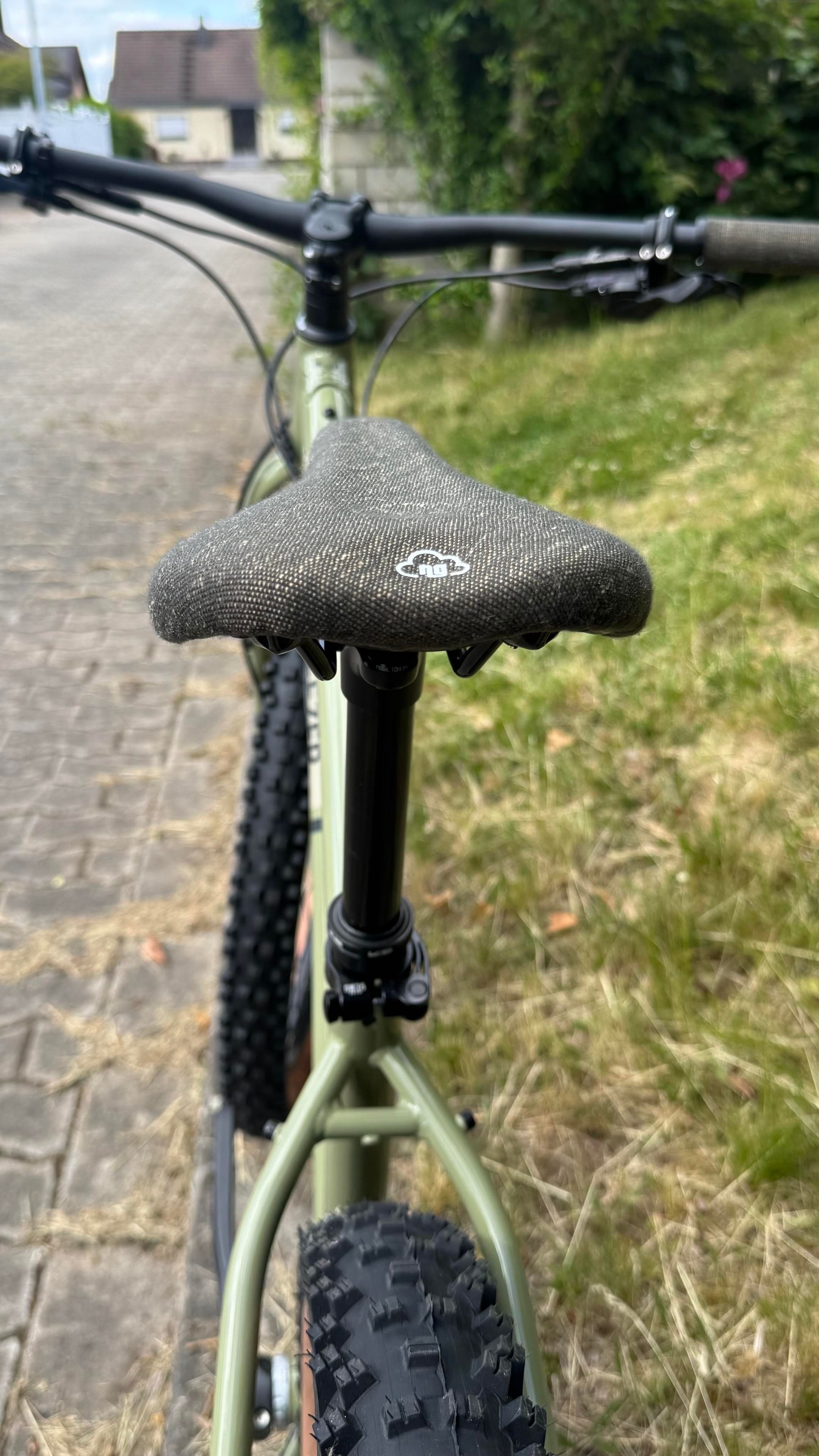 Eine Nahaufnahme eines Fahrradsattels und eines Rahmens. Das Fahrrad verfügt über einen strukturierten grauen Sattel und eine schwarze Sattelstütze. Der Hintergrund umfasst einen Bürgersteigweg und einige Grasflächen.