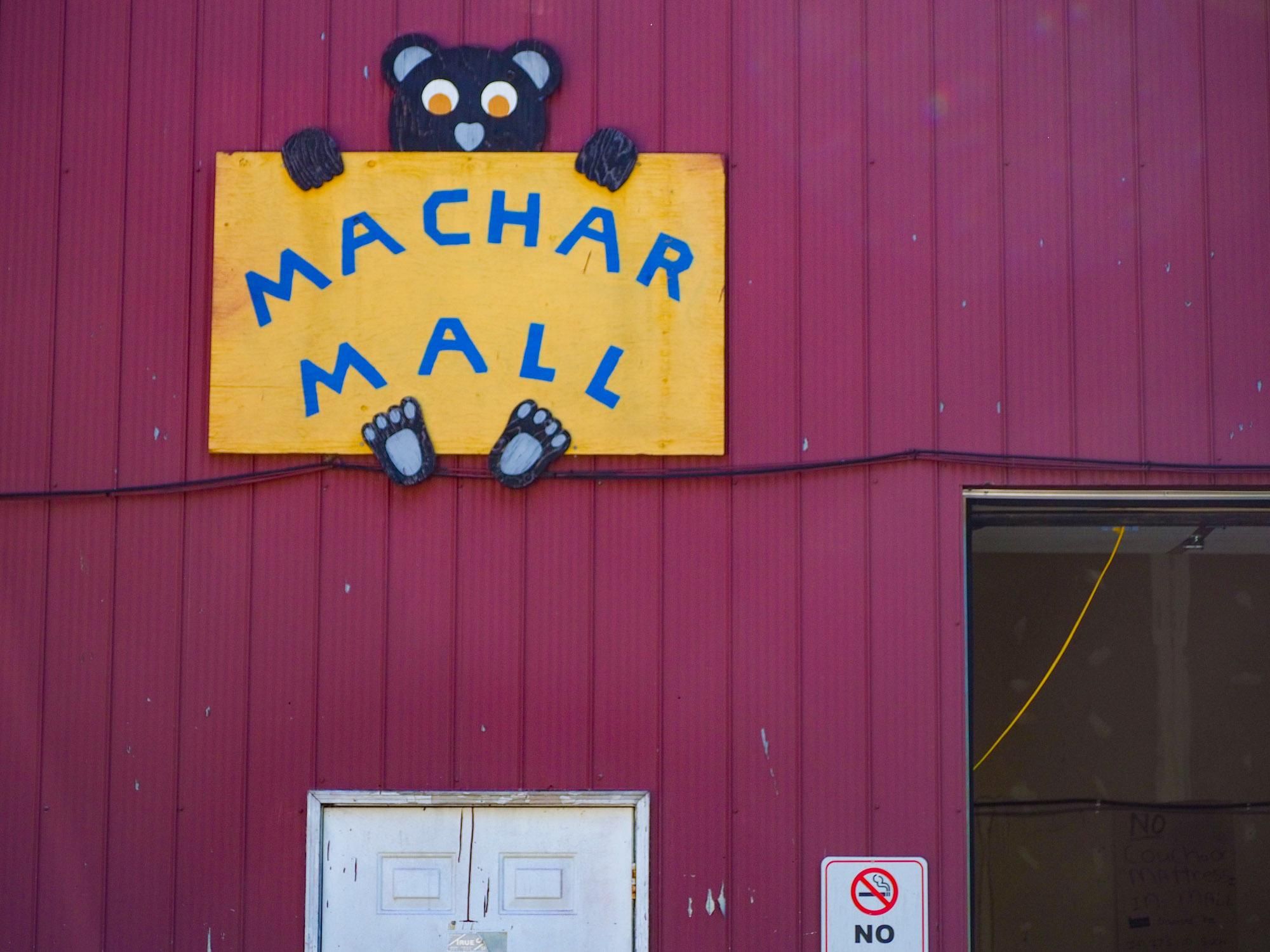 A sign on a red building of a cartoon black bear holding a yellow board with Machar Mall painted on it with blue paint.
