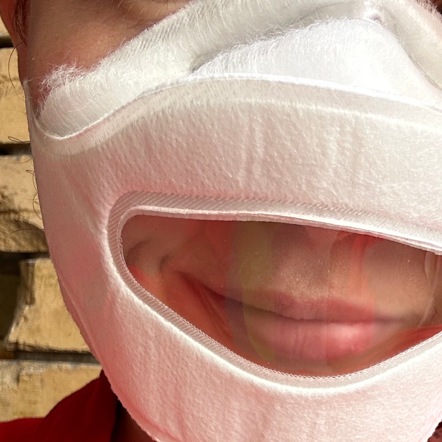 Close-up einer weißen Maske, durch deren Kunststoff-Fenster mein Lächeln zu sehen ist.