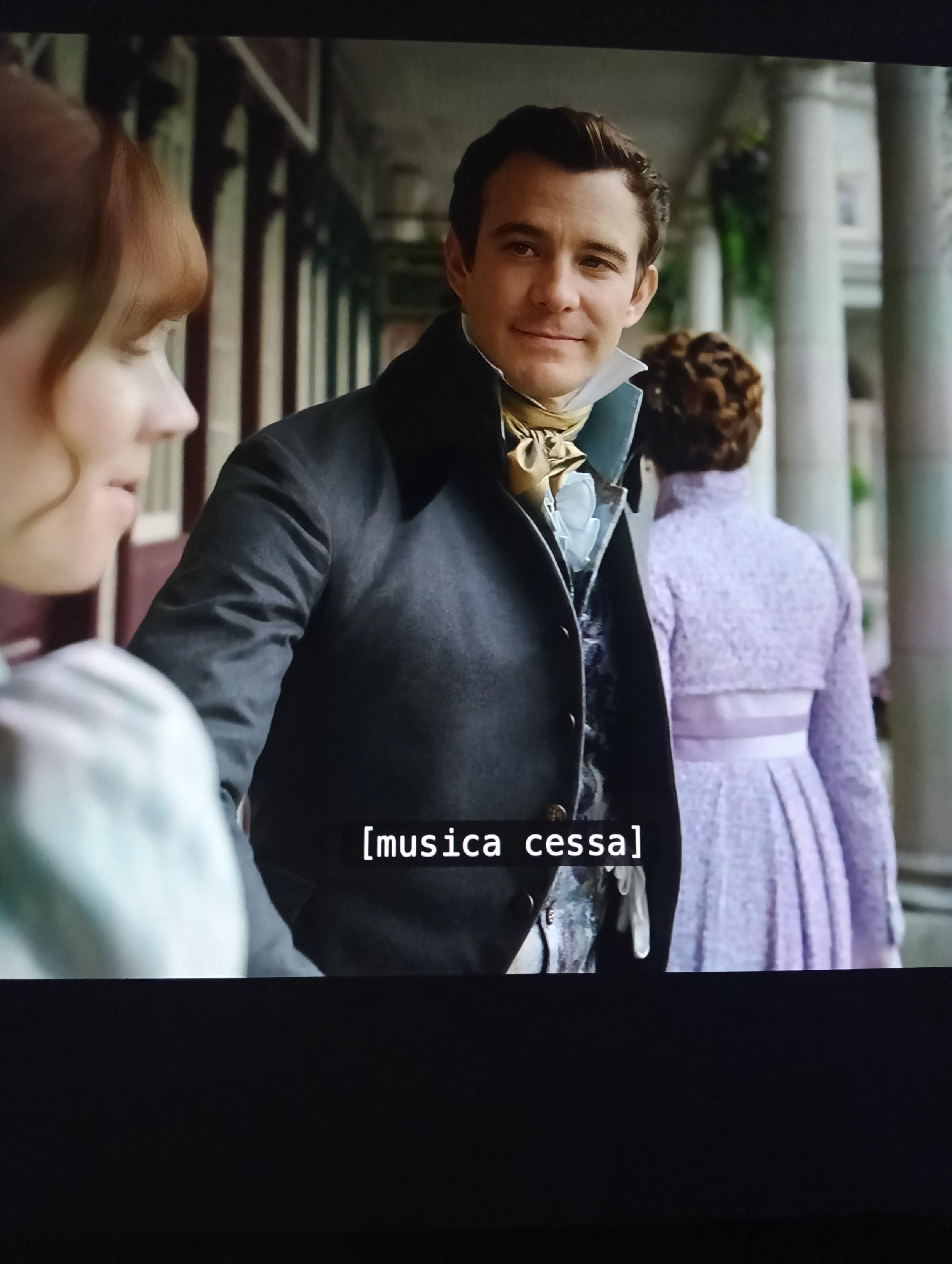 A Scene of Bridgerton.
A well-dressed man in a vintage suit smiles warmly at a woman with auburn hair in period clothing. They stand on a columned veranda. Caption: "[music stops]".