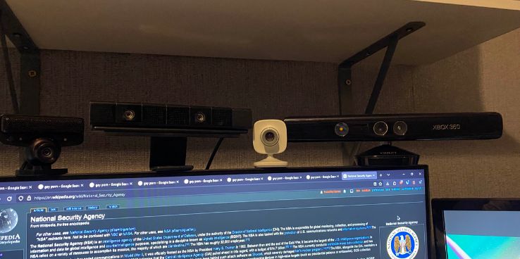picture of the top of my monitor, on top of the monitor are 4 cameras from video game consoles - the playstation 3 eye, the playstation 4 camera, the xbox 360 vision camera and the xbox 360 kinect. all are facing directly towards the viewer. on the monitor itself, the wikipedia page for the national security agency is displayed
