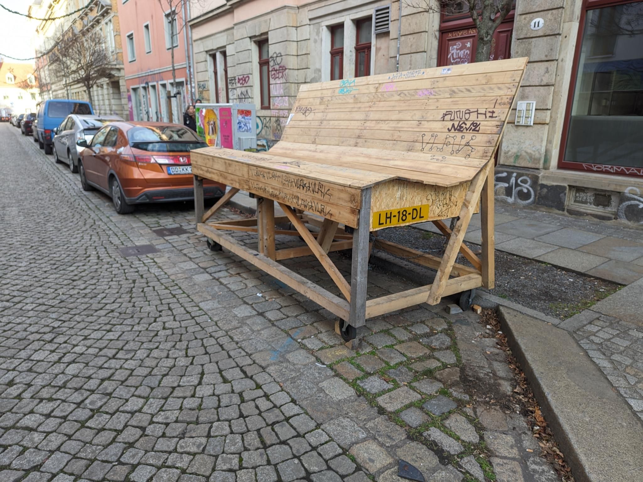 Auf einen Parkstreifen in der Innenstadt steht eine überdimensionierte Holzbank auf Rädern. Es ist ein gelbes (britisches?) KFZ Kennzeichen angeschraubt.
