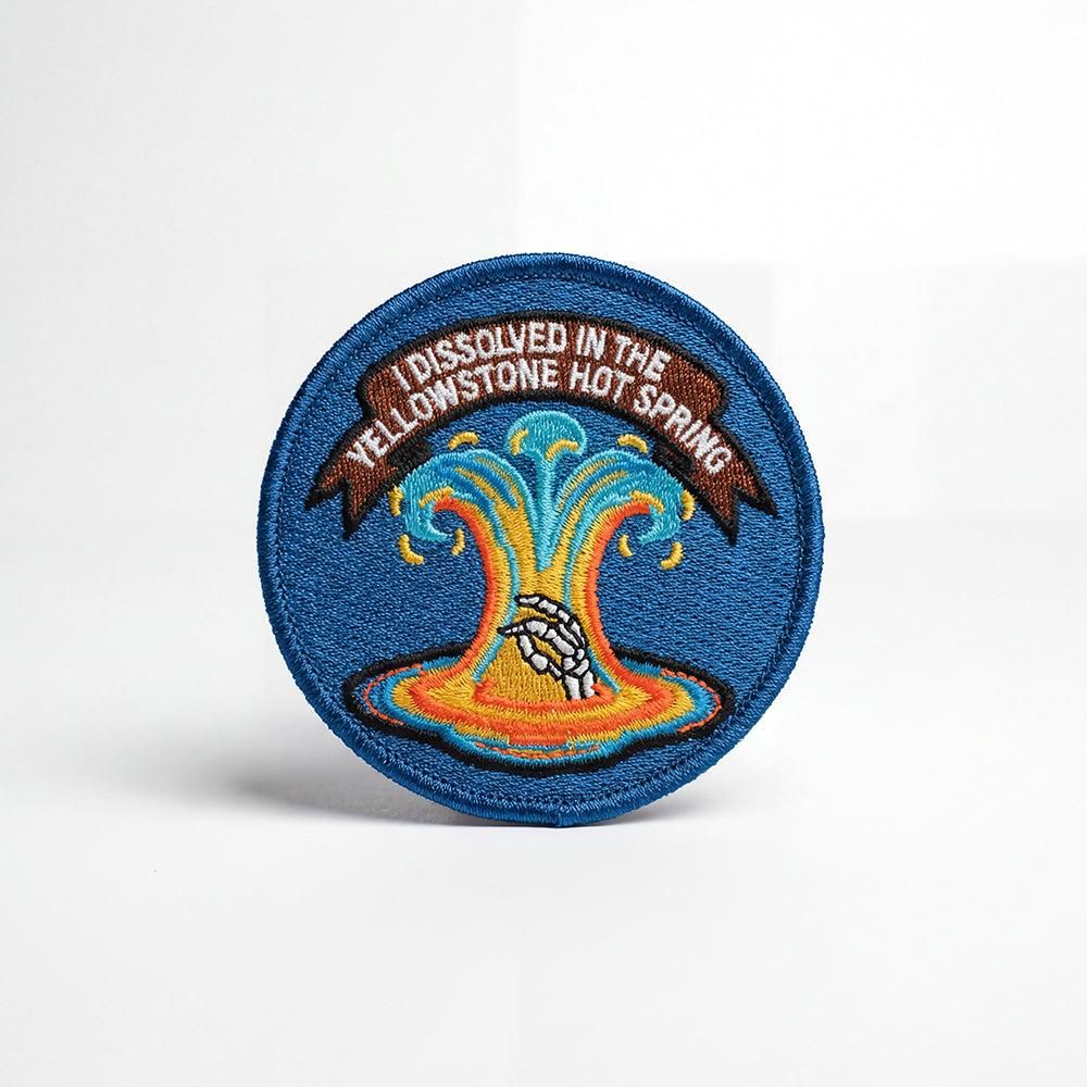 A circular embroidered patch with a blue background and border. At the center, a skeletal hand reaches out from a colorful, steaming hot spring depicted in shades of orange, yellow, and blue. A brown banner above the spring reads, "I DISSOLVED IN THE YELLOWSTONE HOT SPRING" in white capital letters.