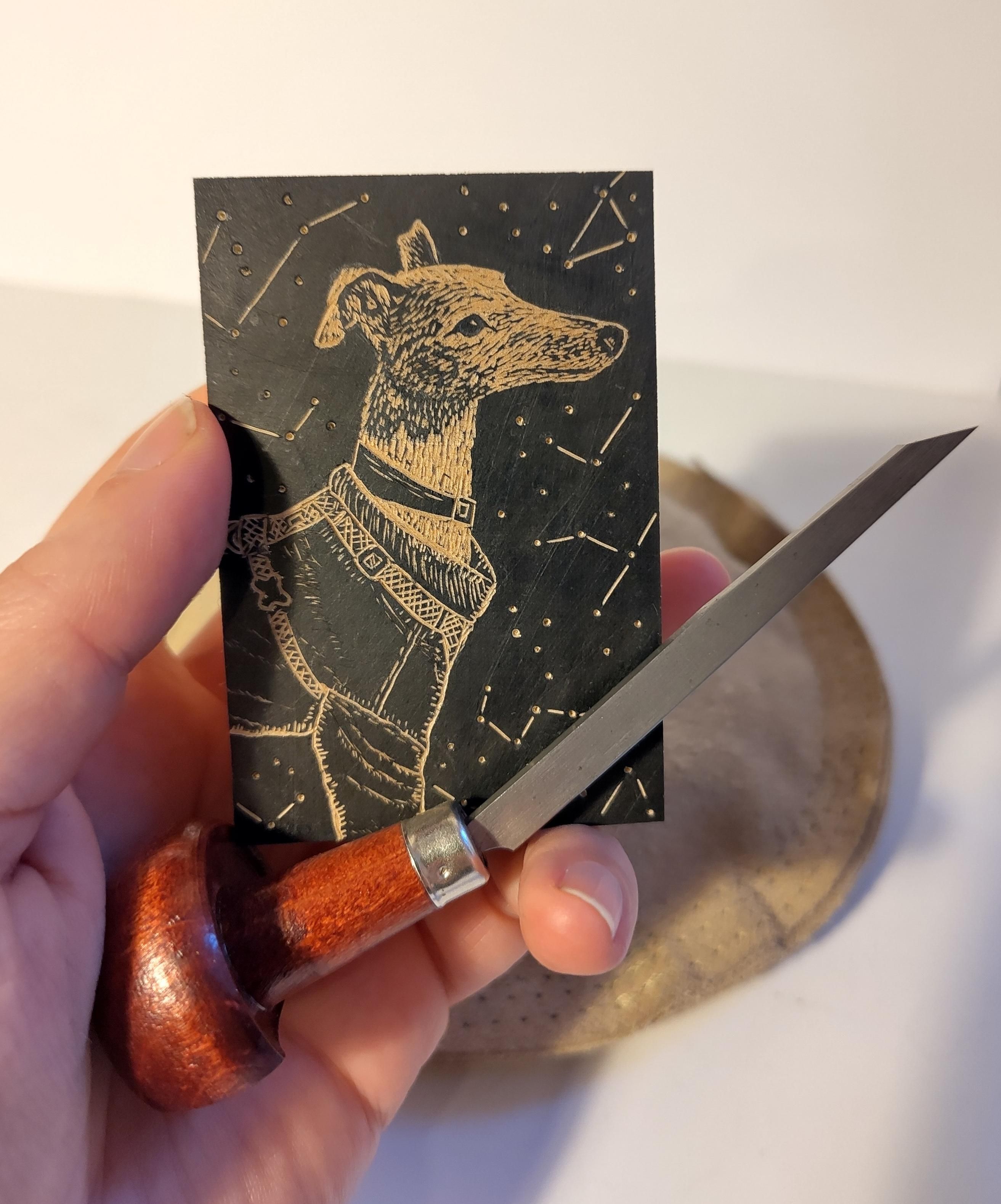 Small wood block held in a hand with an engraving tool. The surface is stained black so the carved bits show up wood coloured, it depicts a whippet in his collared coat, harness and collar, with constellations around him.