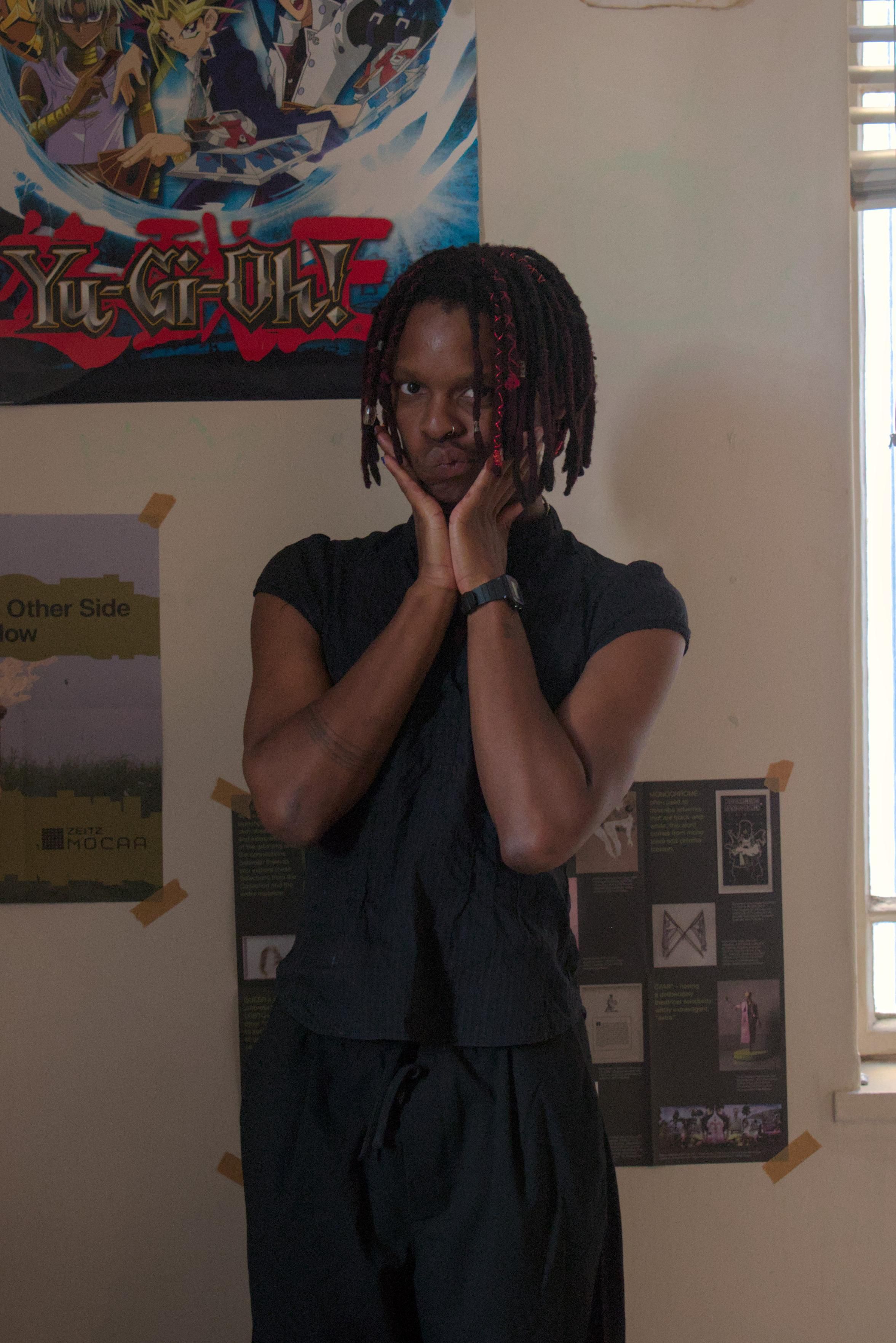 a photo of me in an all black outfit doing a cutesy pose with my hands by my face. A yu-gi-oh poster is in the background.
