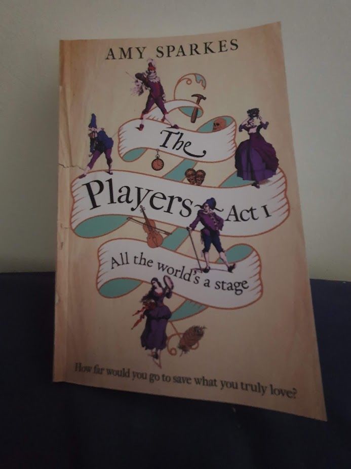 Photo of a book sitting on a blue pillow, propped against a wall. The book is The Players - Act 1, All the world's a stage by Amy Sparkes. 