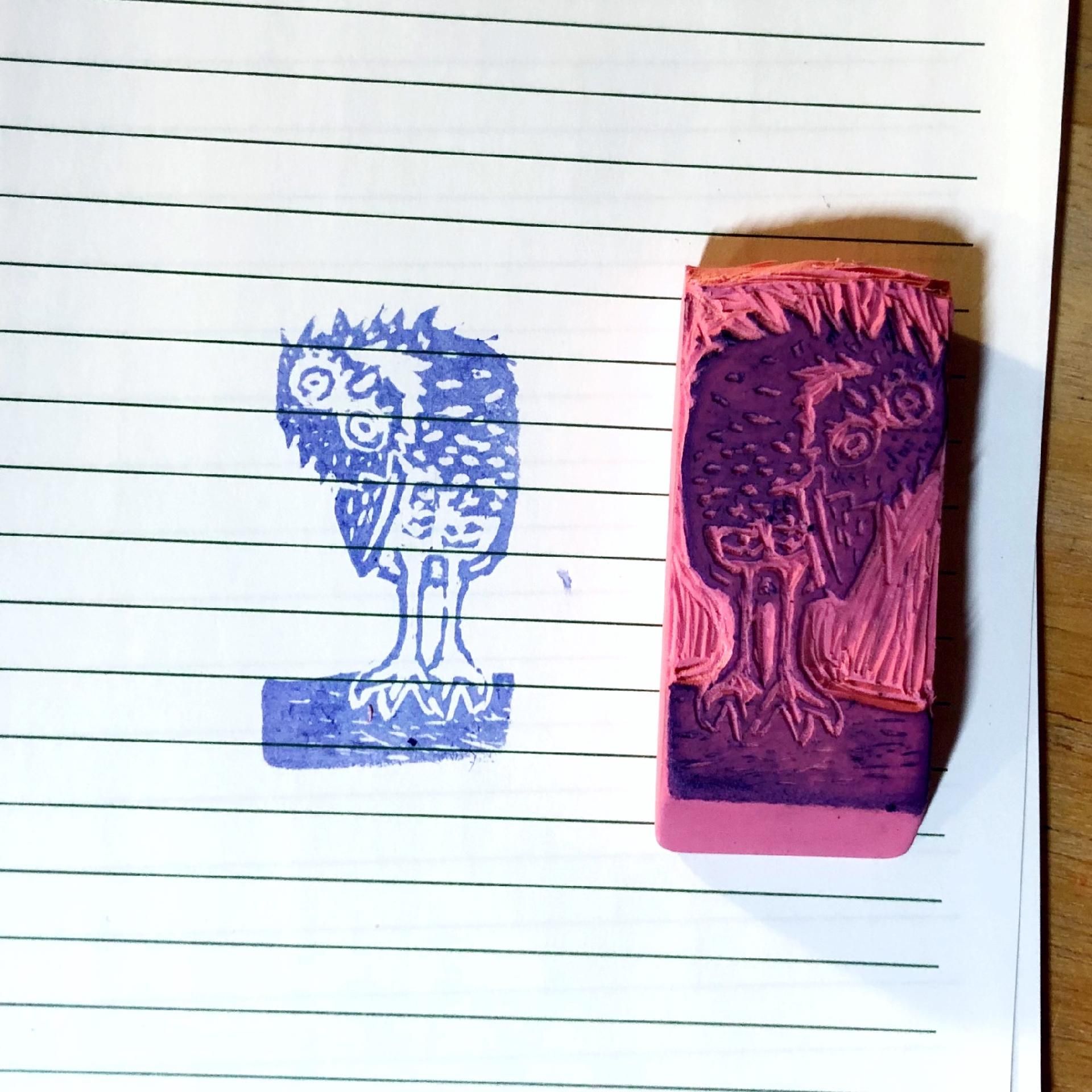 Photo of a piece of note paper with a stamped image on it and a hand carved eraser rubber stamp set on it face up.
The image is of a small burrowing owl with its head tipped over 90 degrees sideways like an extreme confused dog head tilt