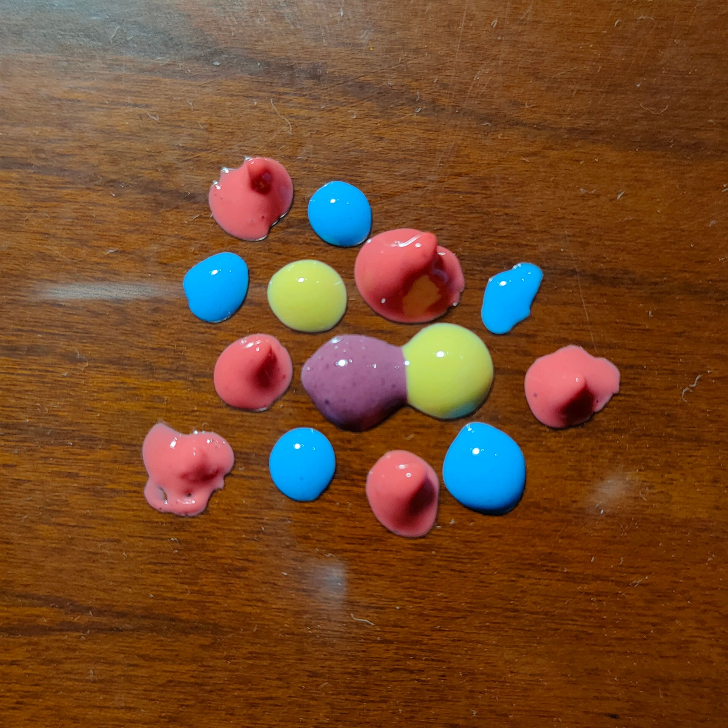 Several drops of badly mixed paint over a sheet of plastic on a wooden table. The drops are close together. They're pink, blue, yellow and purple, because I had those colours for some other project.