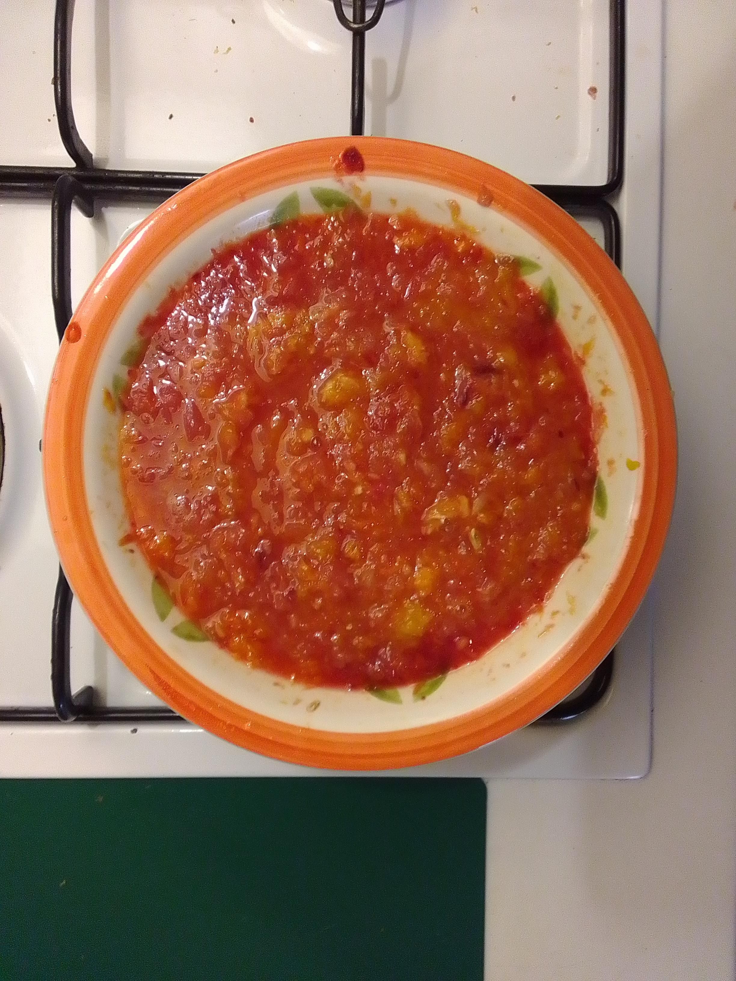 Foto a colori in formato verticale che ritrae dall'alto un piatto fondo bianco e rosso poggiato su un fornello e colmo di residui di succo di arance e pompelmi, un liquido un po' grumoso di colore rosso con parti arancioni.