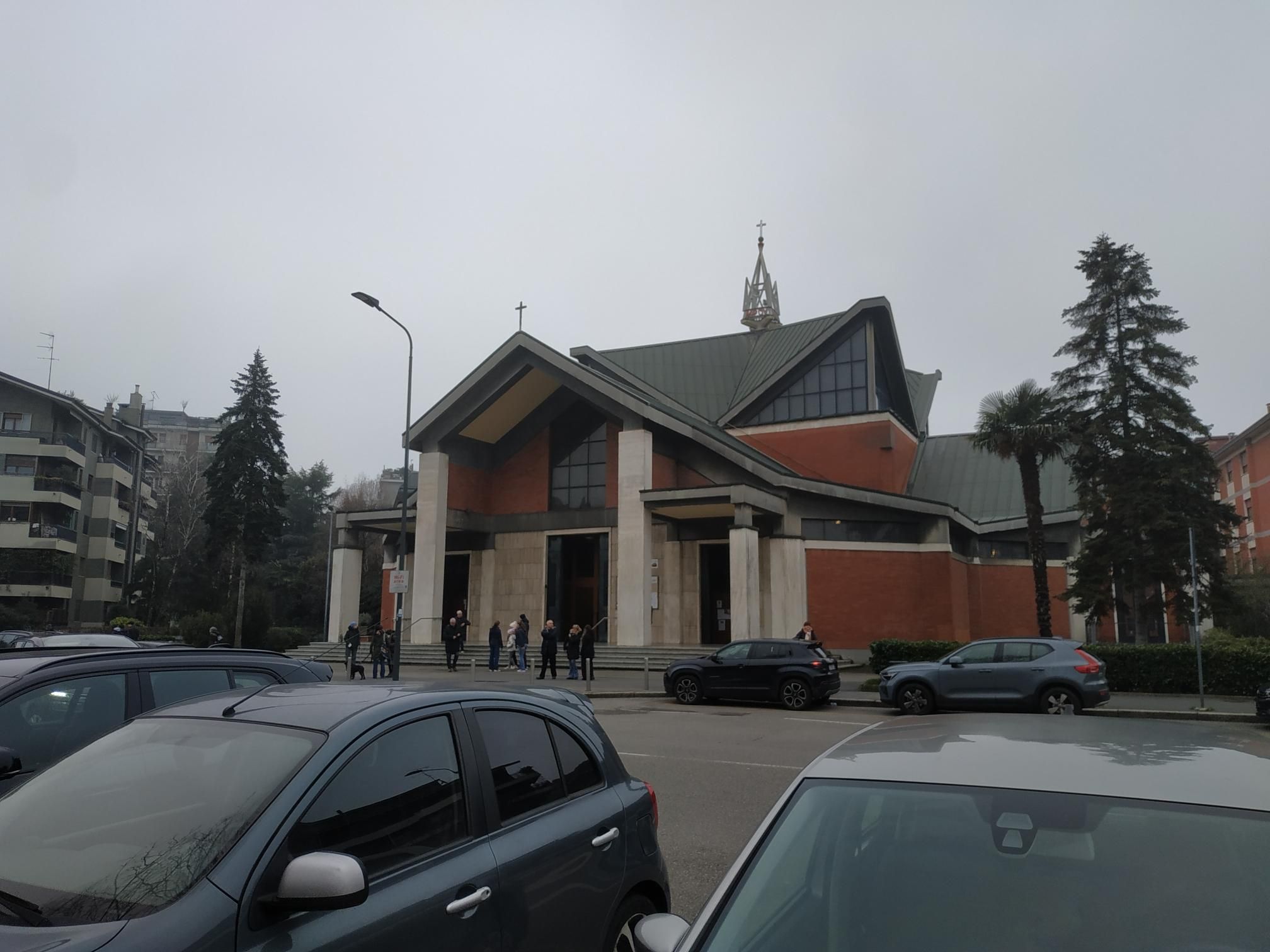 stessa chiesa dove si vede meglio la facciata con un ingresso alto tra due colonne e la gente che esce dalla funzione religiosa. in primo piano delle automobili.