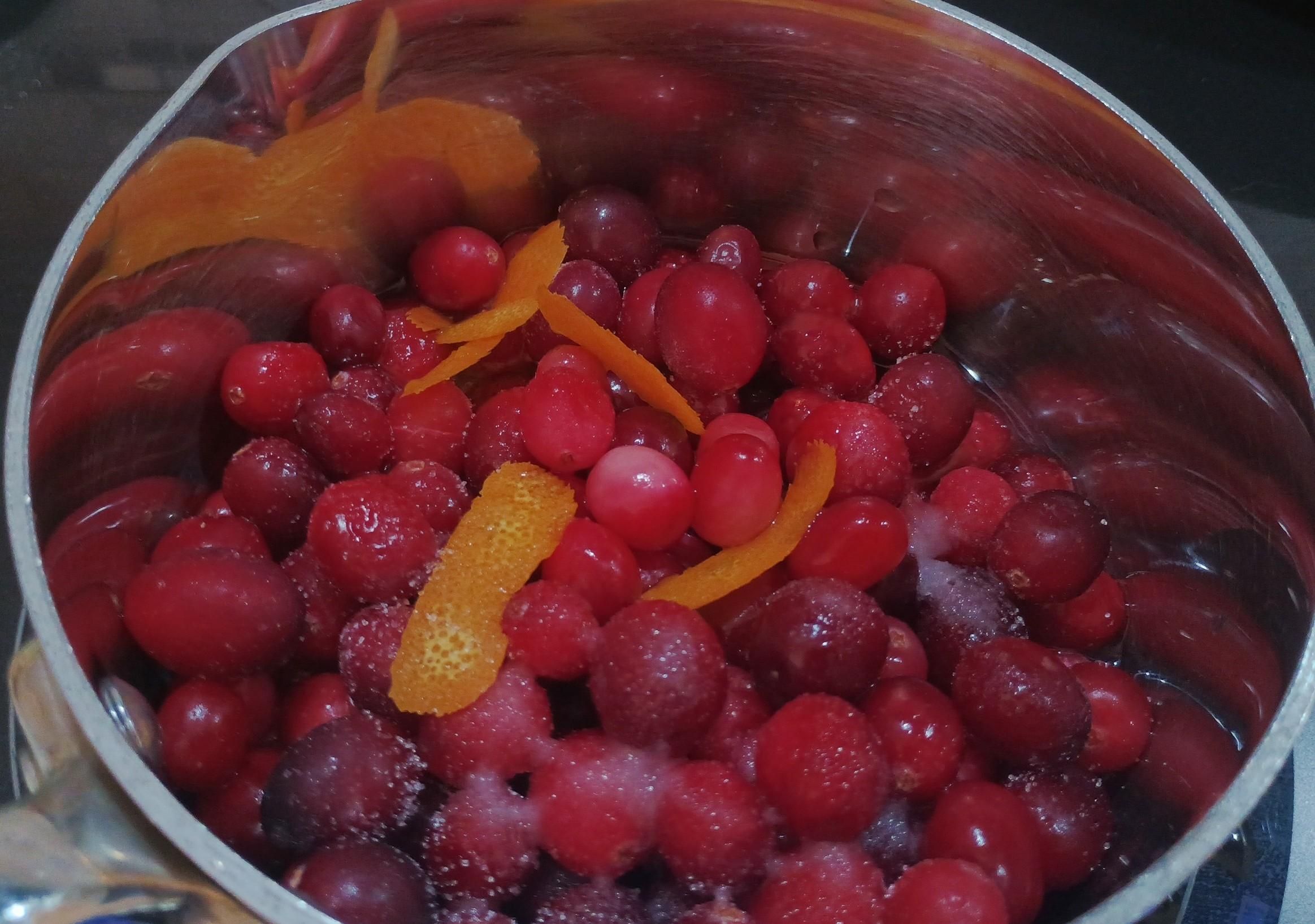 A saucepan on the stove contains red cranberries, orange peel and some sugar, just before it gets heated up for sauce making.
