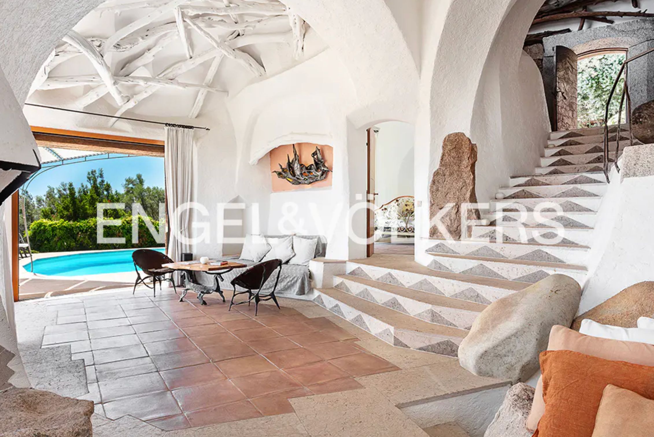 A large open room with curved walls and part of the bowed ceiling has cris-crossing painted white sticks attached to it. The floor is terracotta coloured tiles, and there is a stairway with large rocks scattered around it. 