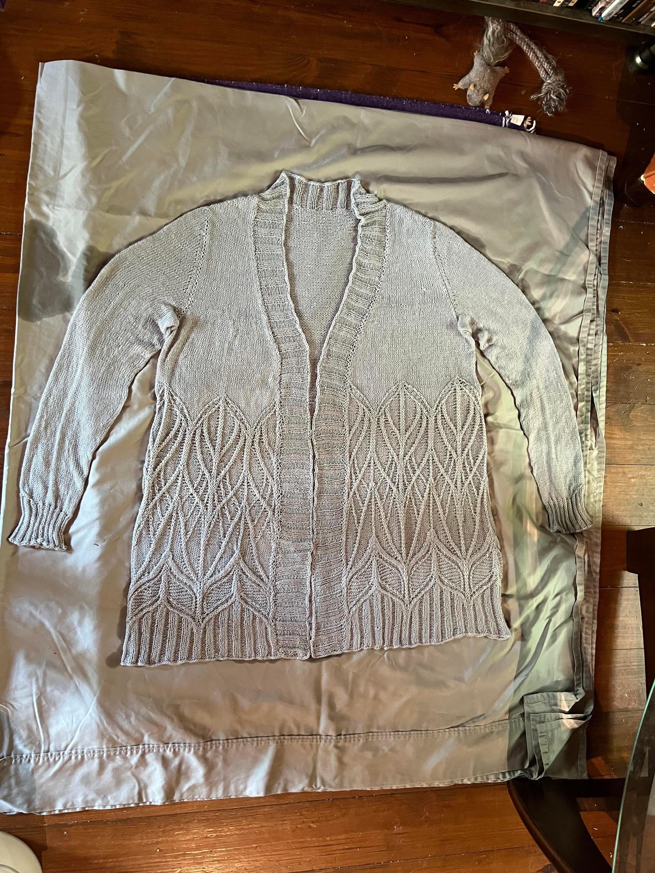 Grey sweater with a fine tracery of cables on the bottom part blocking on a greenish sheet.