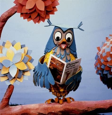 Animated owl -doll ; blue with yellow, holding a newspaper. Standing on a tree-branch. Colorful animated branches on both sides.