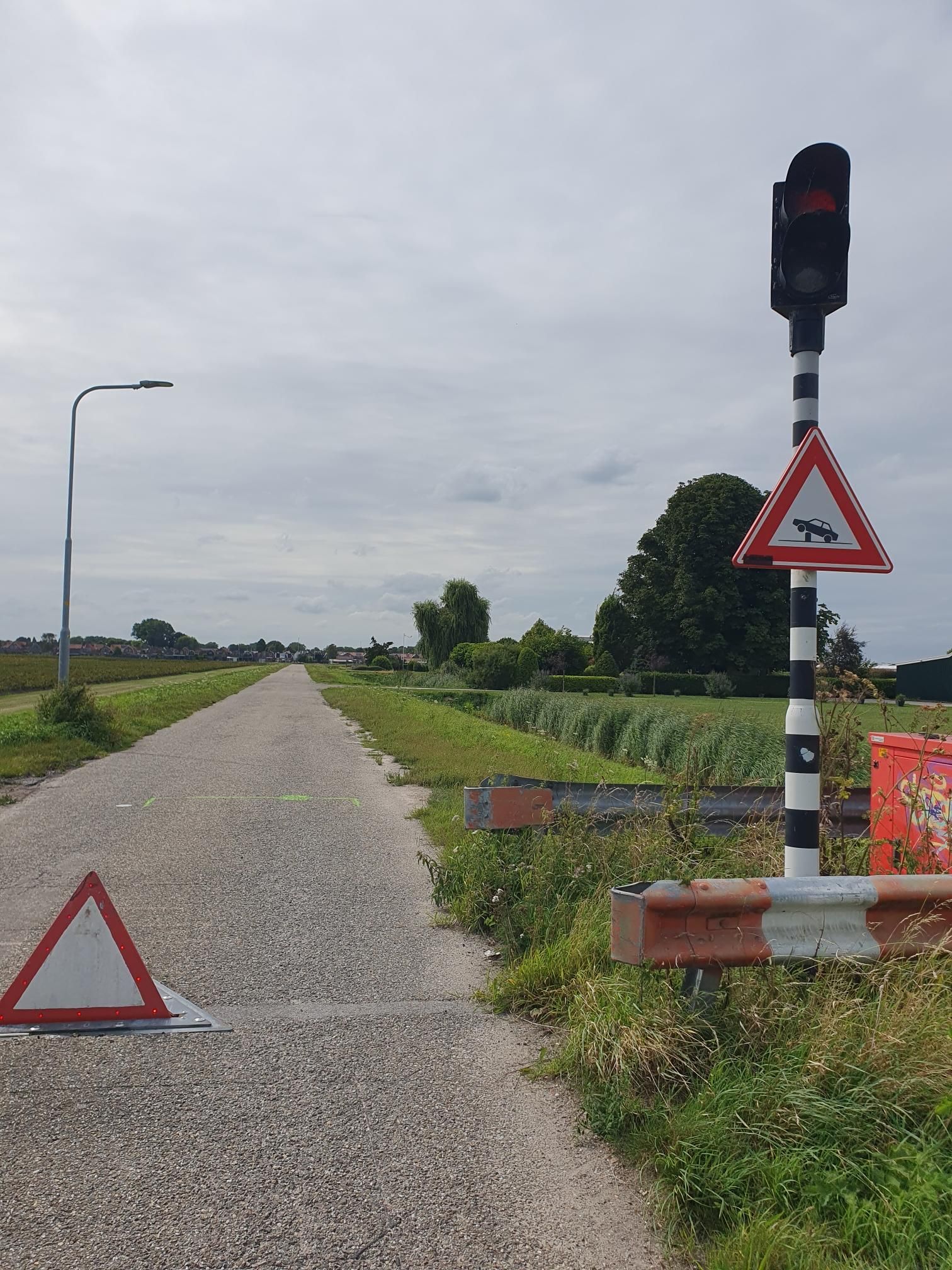A small road (or maybe a bike path, who knows) flanked by grass, with a red traffic light on the right, and a warning sign below it depicting a car stuck on a bollard. In the middle of the way there is a triangular thing (like the warning sign, but without the symbol) sticking up from the ground.