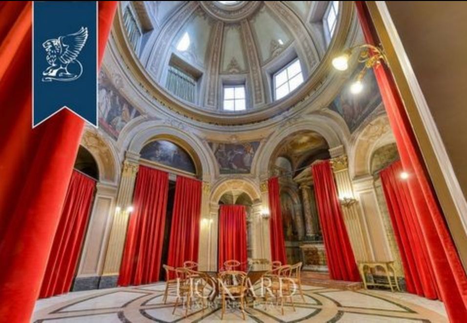A huge round room with giant domed ceiling and arched doorways all around it, painted with murals above the doors, and the dome has windows and additional painted adornment. There are red curtains on each of the doors, and the floor is marble with a circular design. 