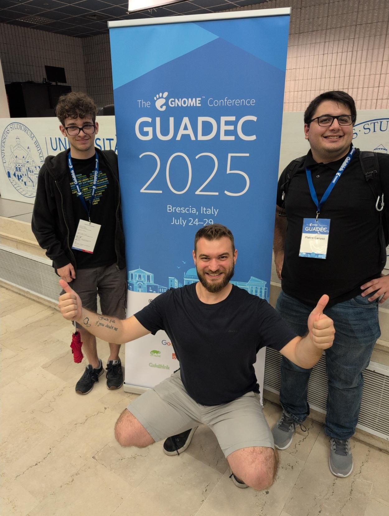 Photo of me and ITzTravelInTime with Pietro di Caprio around the GUADEC 2025 banner.