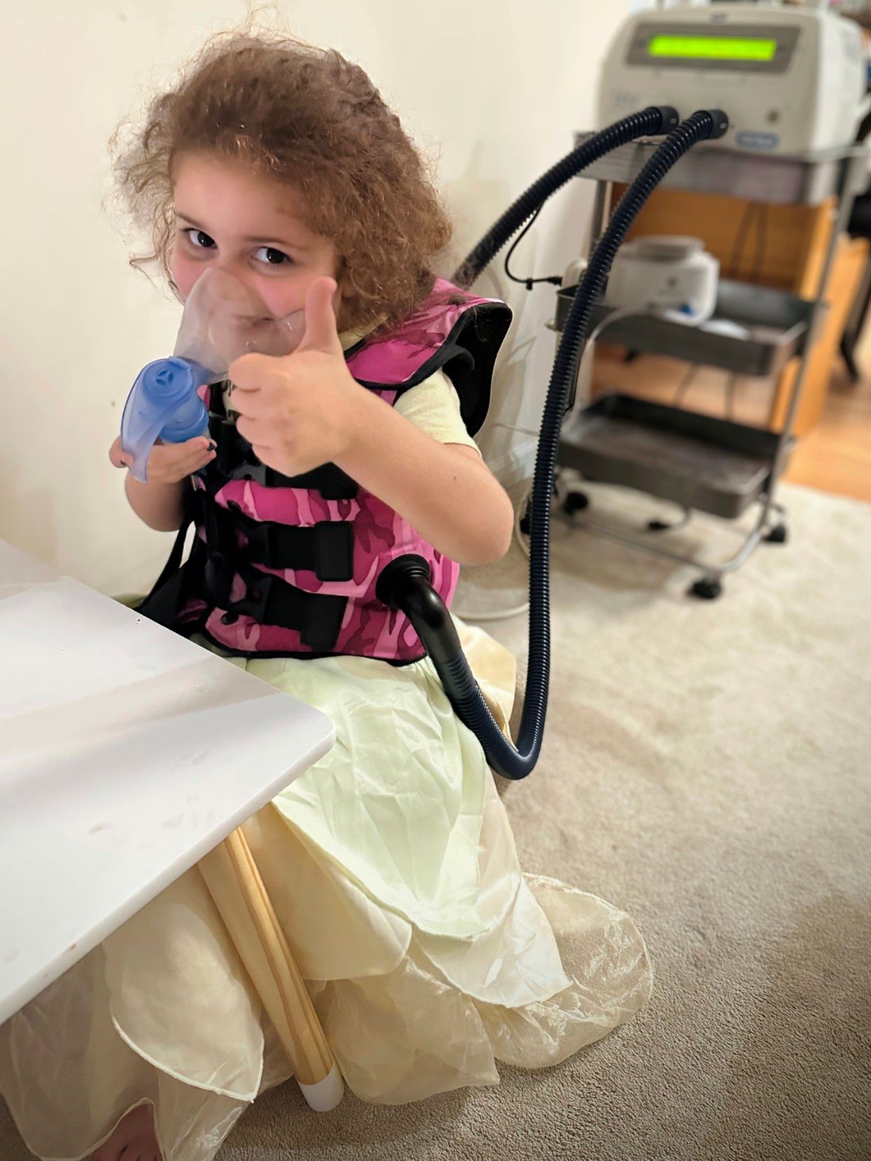 Toddler smiling to the camera and giving a thumbs up while wearing a high-frequency chest wall oscillation vest and using her nebulizer to deliver medications to her lungs.
In addition to medications like Trikafka and enzymes with every meal & snack to help her metabolize food, this is a twice daily treatment for a total of about 1½ hours.