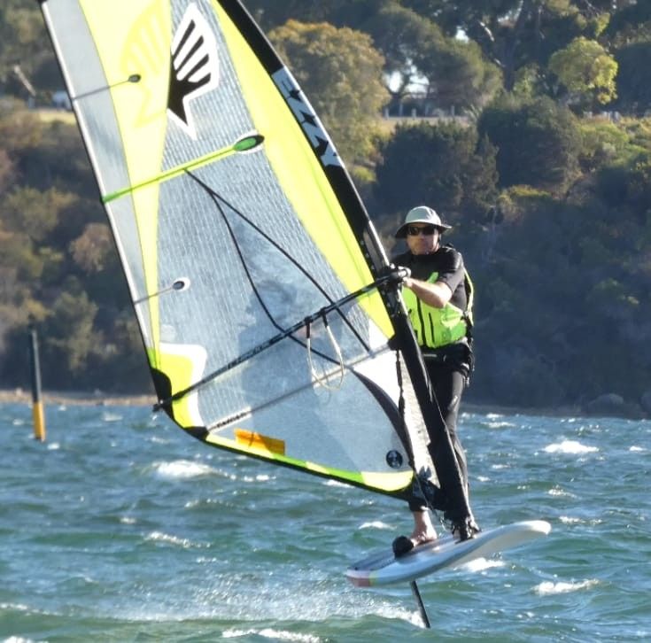 Wind foiler with yellow sail and life jacket coming towards you starting to turn.