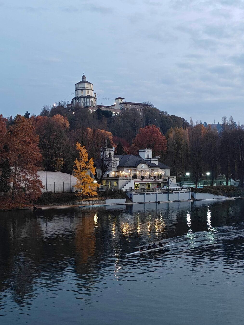 View of the Po River in Turin, Italy, with a rowing shell in the foreground, a brightly lit club building on the bank, and the large white dome of the Monte dei Cappuccini church atop a tree-covered hill in the background under a twilight sky.