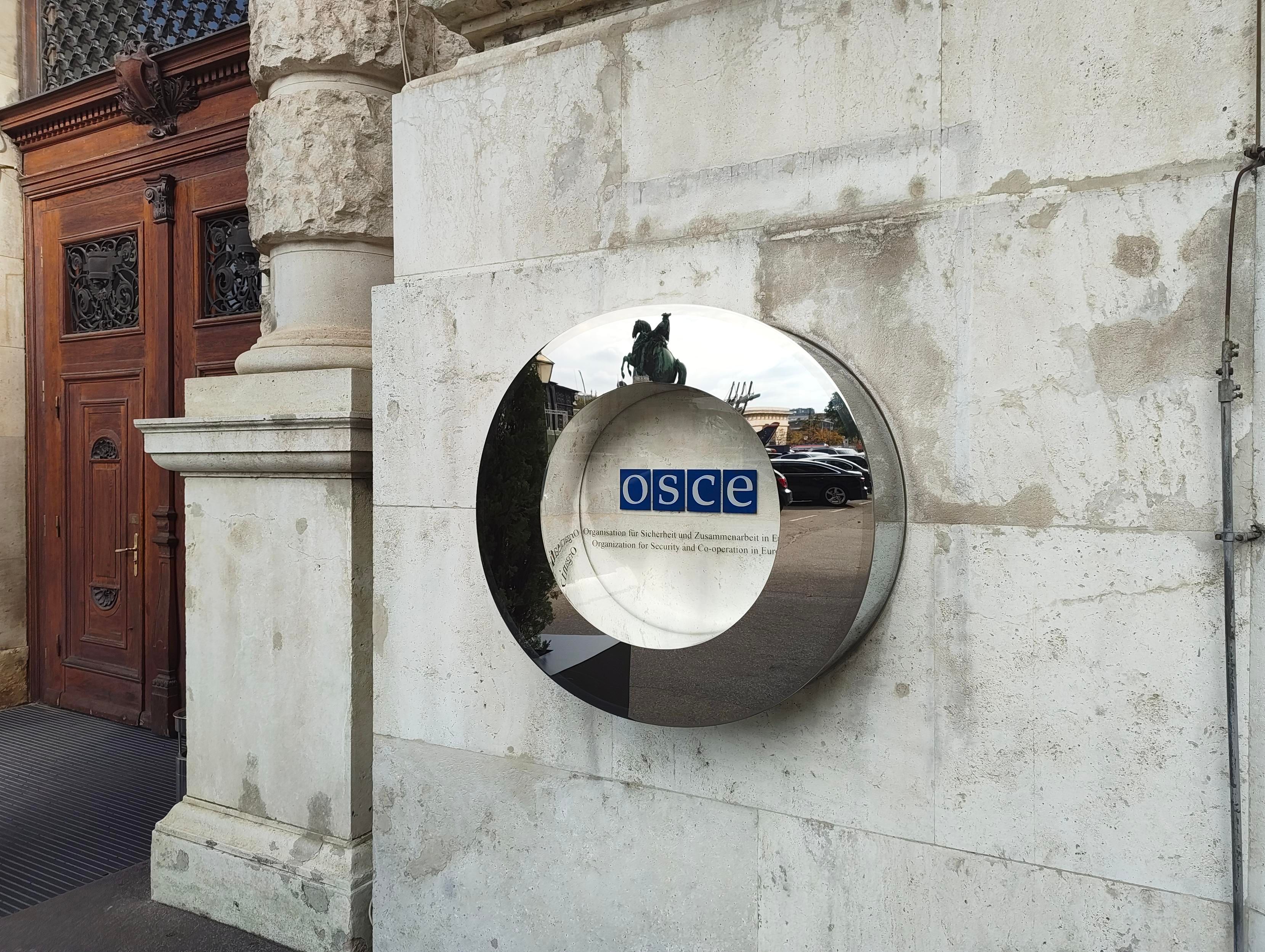 OSCE logo on a grey stone wall of Hofburg with a round mirror as decoration where the statue of Archduke Carl on the Heldenplatz in Vienna is visible