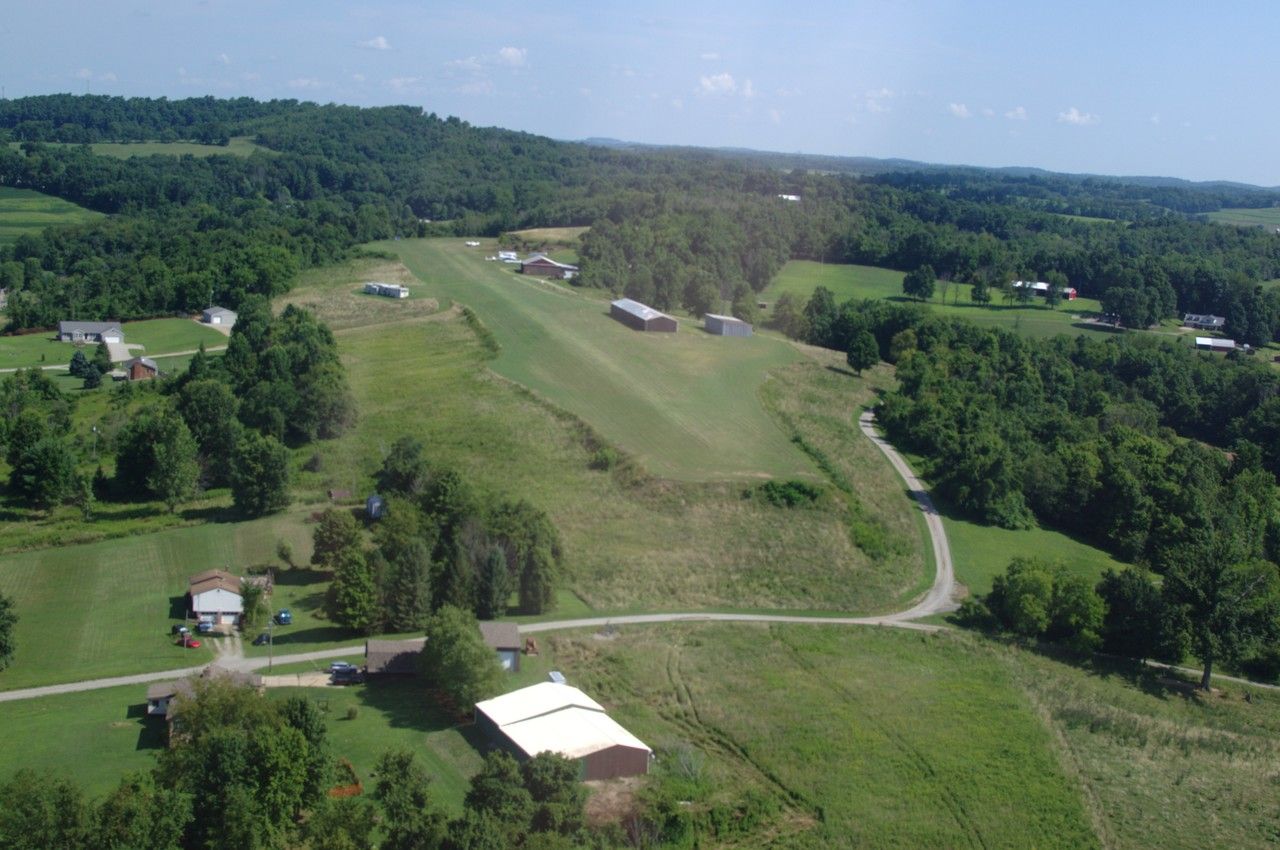 Bendel Airport as seen on the baseleg approach. It's a flattened grass field with a gravel center extending about 3000 feet into the distance. In front of and below the grass/gravel strip is a road. Two red buildings are to the right.