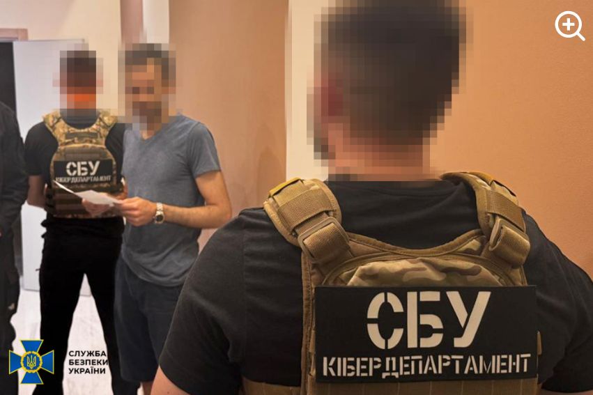 An unnamed 38-year-old man was arrested in Kiev last month on suspicion of administering the cybercrime forum XSS. Image: ssu.gov.ua. He is wearing dark shorts and a blue-grey t-shirt and is holding a document. His face is blurred out, and he is standing between two men in SBU uniforms.