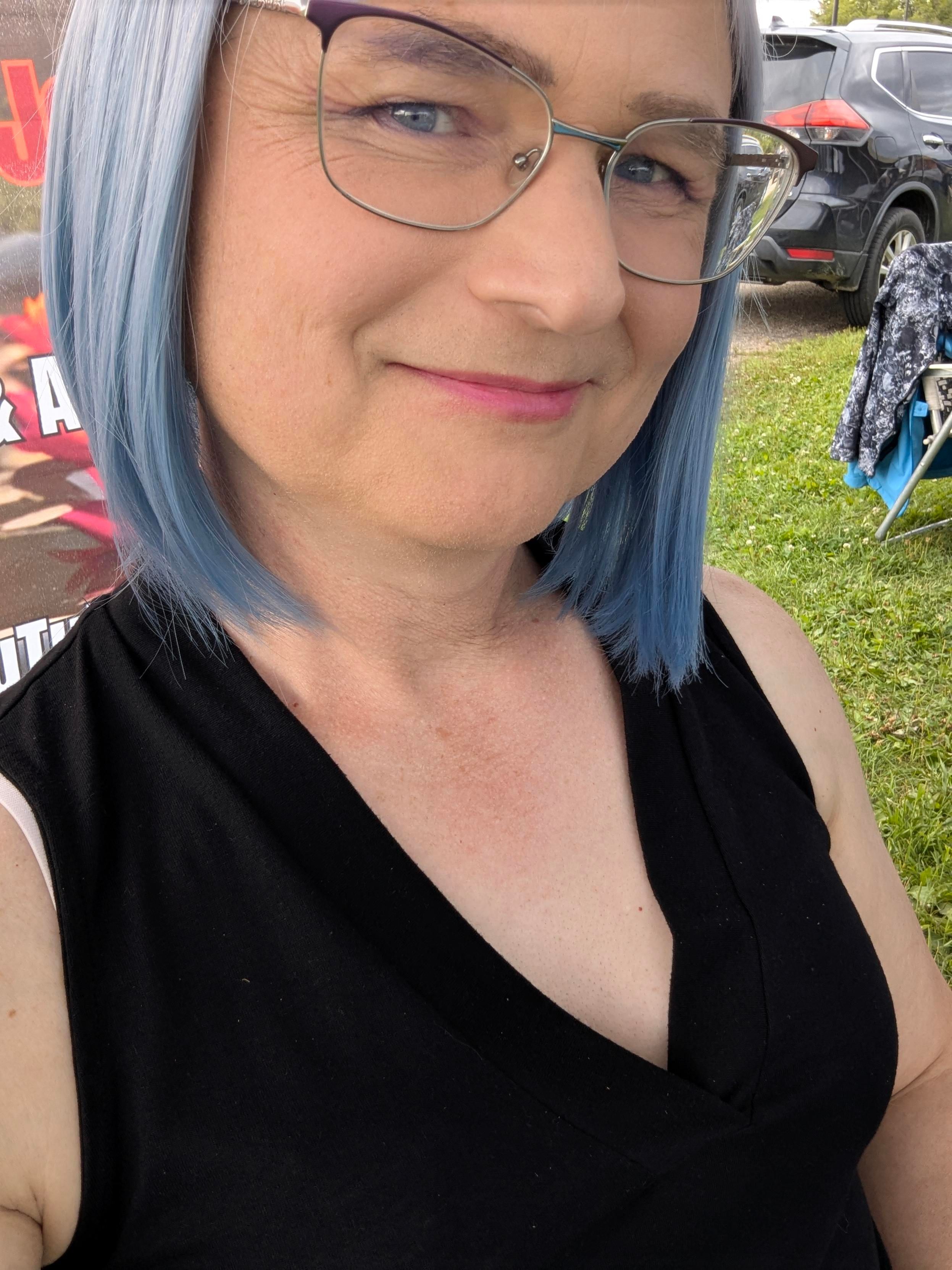 A selfie at an outdoor market. I'm wearing a light blue bob wig and a low v-neck black top over a push up bra that shows a hint of my cleavage.