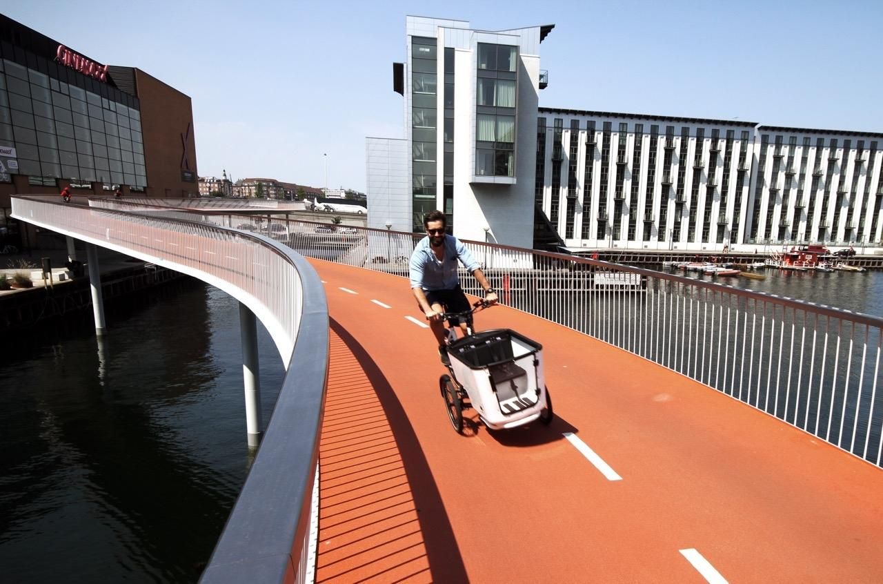 A person drives a cargo bike over a red bike path going over water as a bridge.