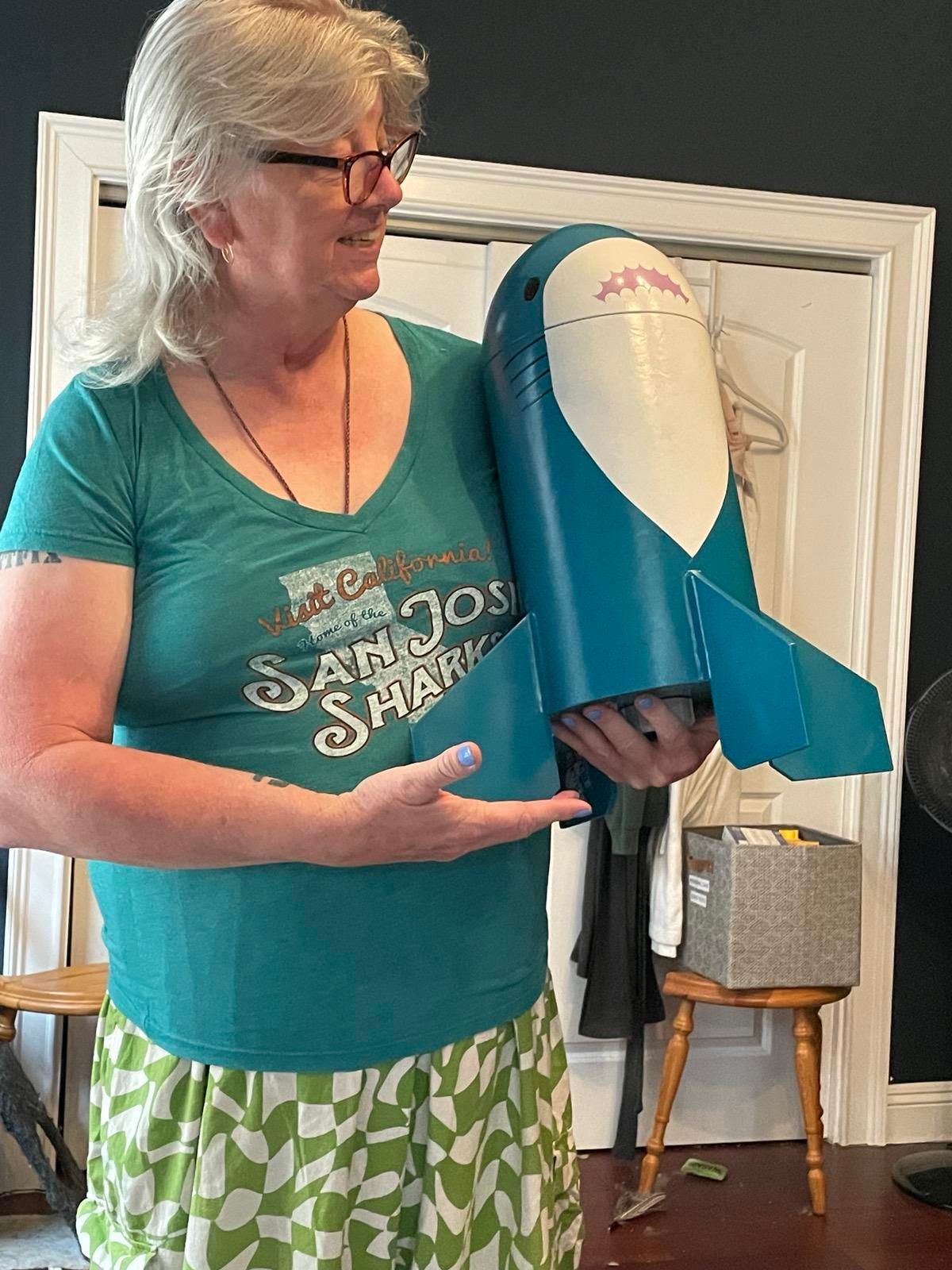 A smiling white woman with glasses holding a model rocket, decorated like a Blåhaj, like a small child 
