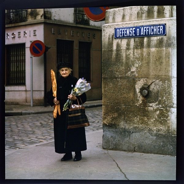 Elderly woman in overcoat and hat on a street corner holding fresh baguettes, a bouquet of flowers, and a hand bag.