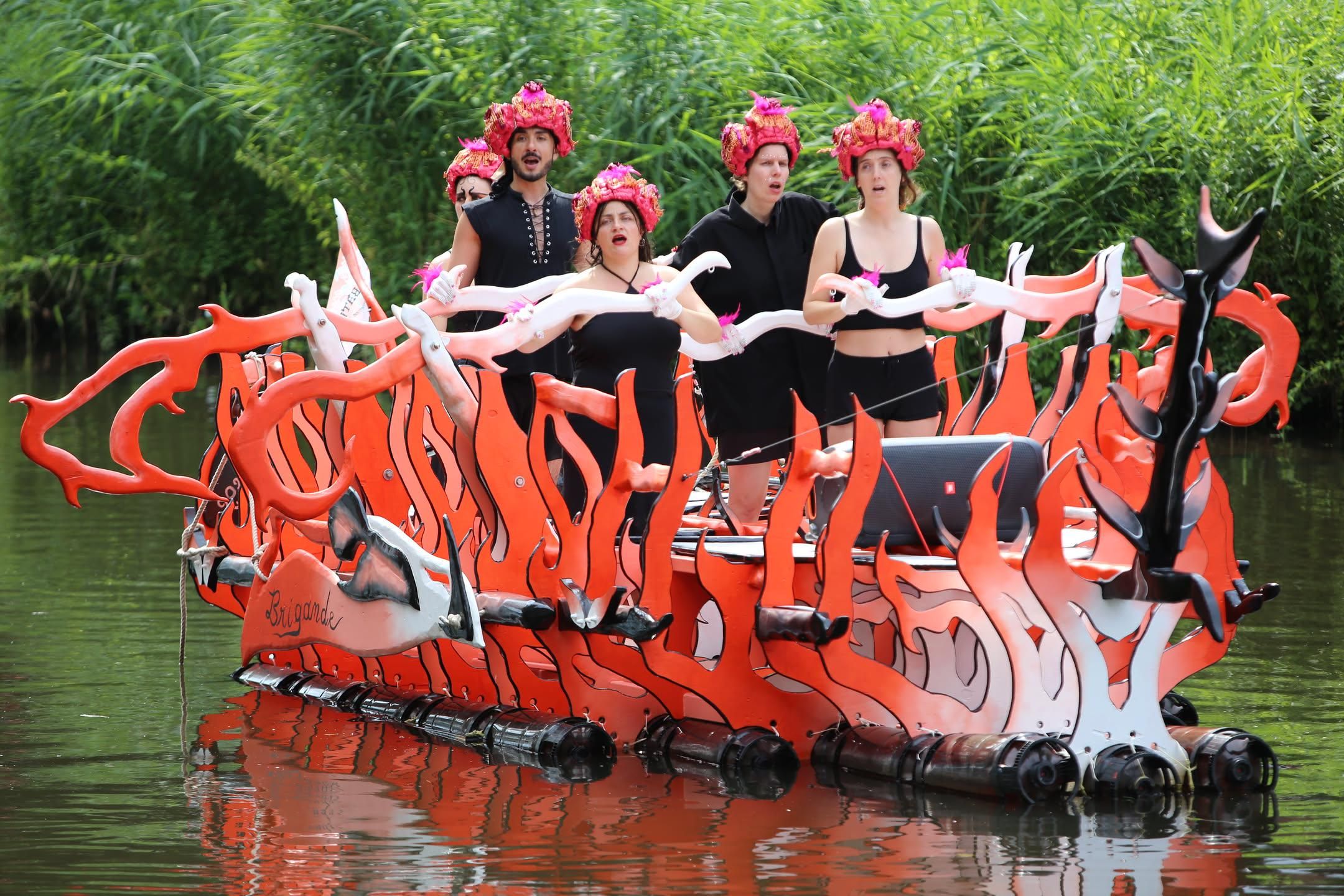 five people dressed like Sirens singing a sea shanty while "rowing" a boat that looks like flames