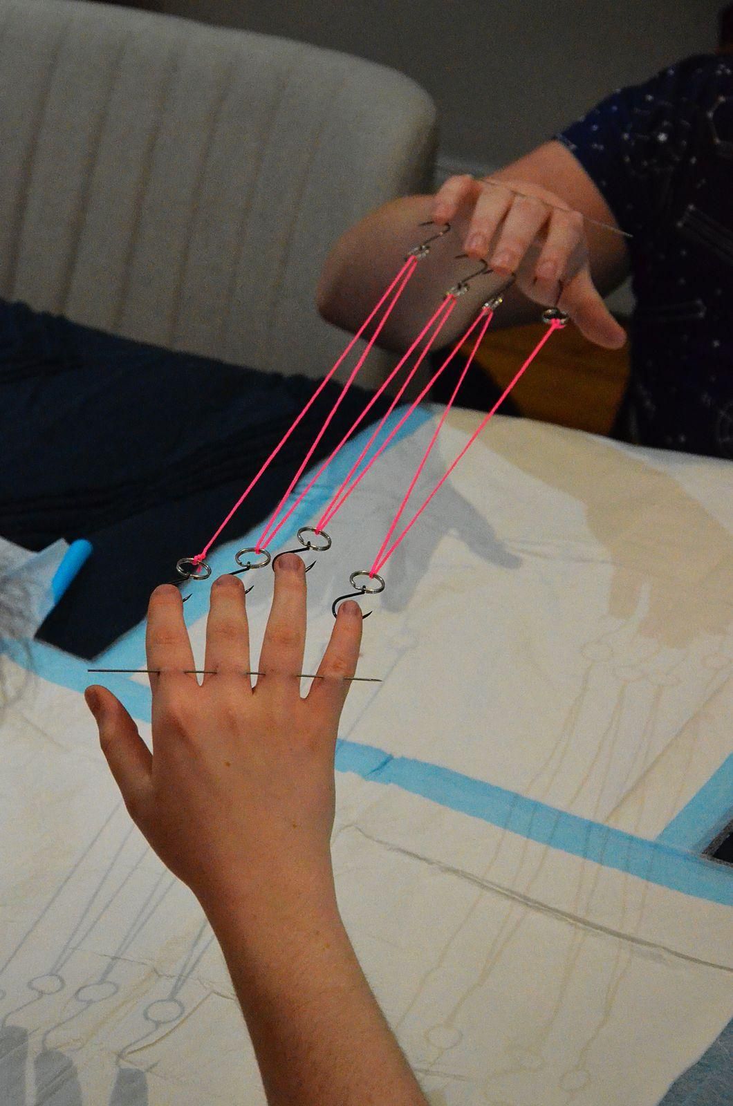Two people hold out their hands. Both have been pierced with hooks through the fingers. The loop attached to the hooks has been threaded with neon pink thread in a criss cross. Any movement on one person transfers mostly evenly to the other. Across the backs of the fingers is a single long piercing needle pinning the knuckles near the base of the finger.
