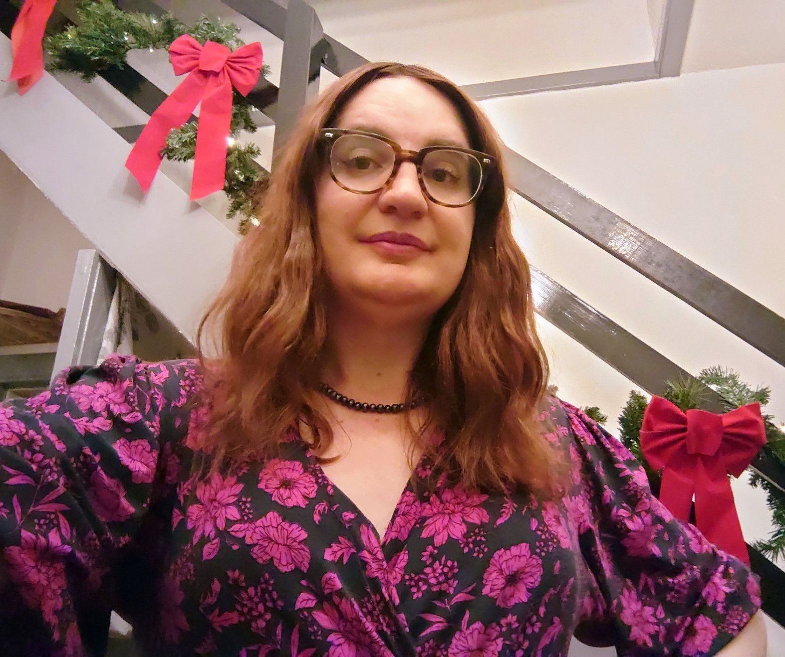 This is me. I'm a non-binary woman with long, wavy chestnut hair, white skin with a warm yellow tint, hazel eyes, and Mediterranean features. In this photo I have my hands on my hips and a gentle smile on my face. I'm wearing round tortoiseshell glasses, a black dress with fuchsia flowers, and a black pearl necklace. I have on mascara, winged eyeliner, and plum lipstick. Behind me are Christmas decorations-- fake pine garlands with huge red ribbons, wound round the stair rails.